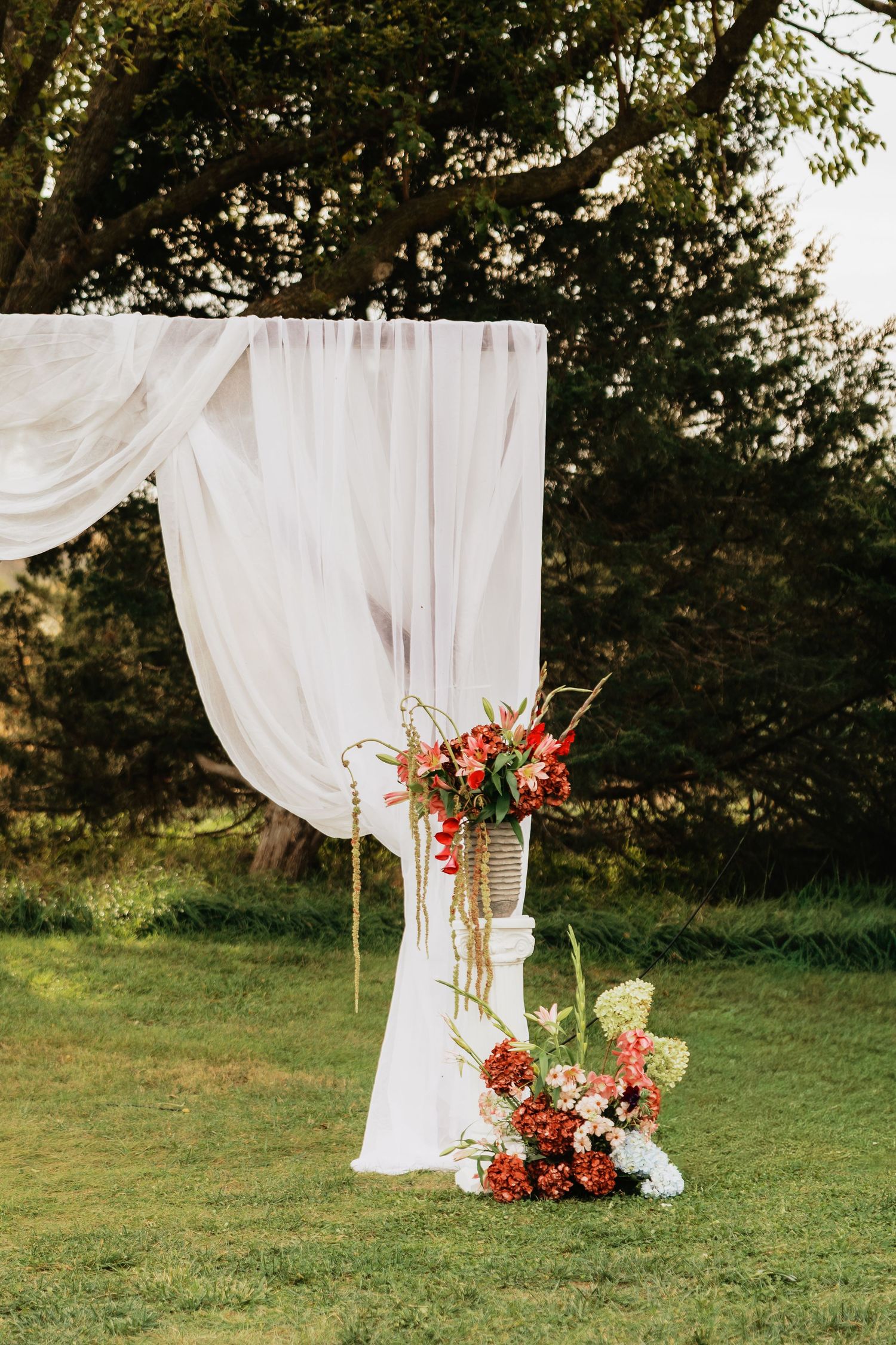 Outdoor wedding ceremony arch draped in flowing white fabric with a red and coral floral arrangement beside it on a green lawn.