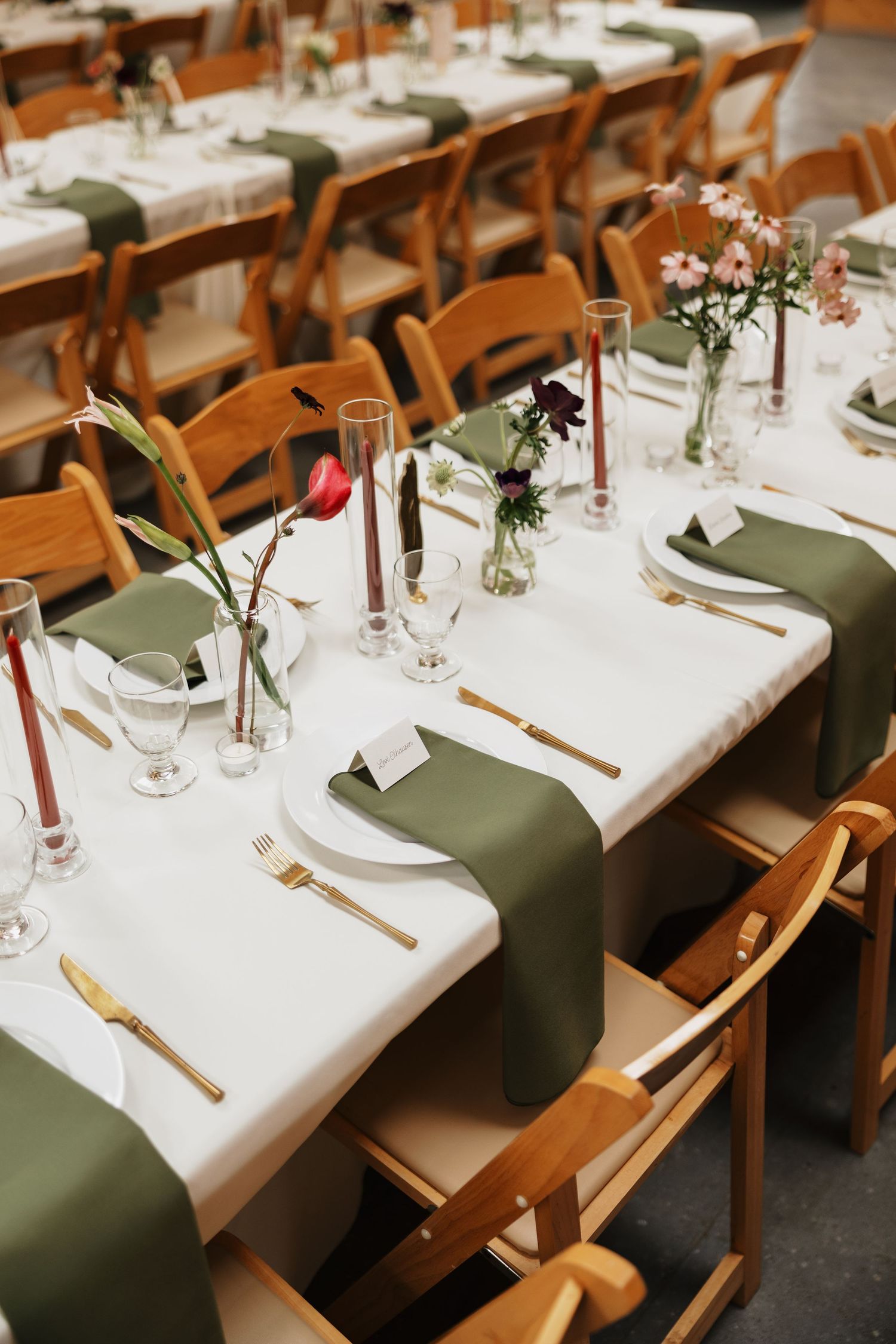 Long wedding reception tables with white linens, olive green napkins, red taper candles, wildflower vases, and wooden folding chairs.