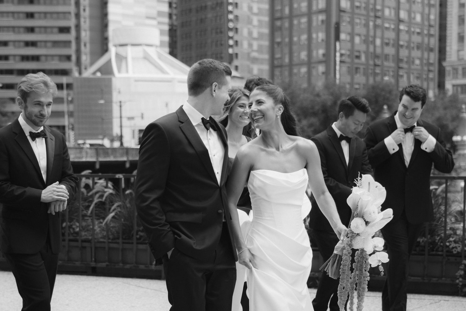 Black and white wedding photo of bride and groom sharing a moment with groomsmen on an urban rooftop.