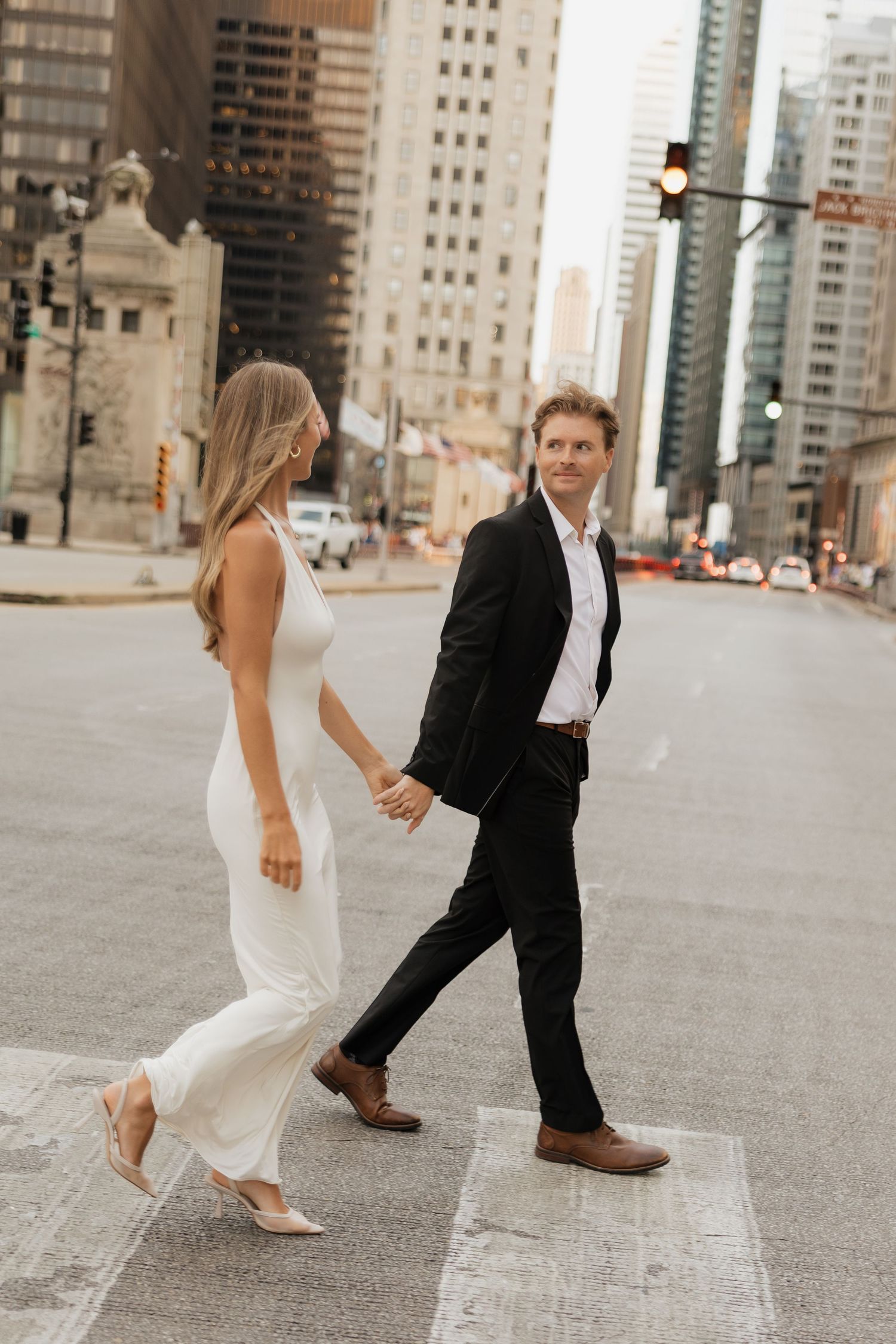 Elegant couple holding hands crossing a city street, woman in white dress, man in black suit, urban skyline backdrop.