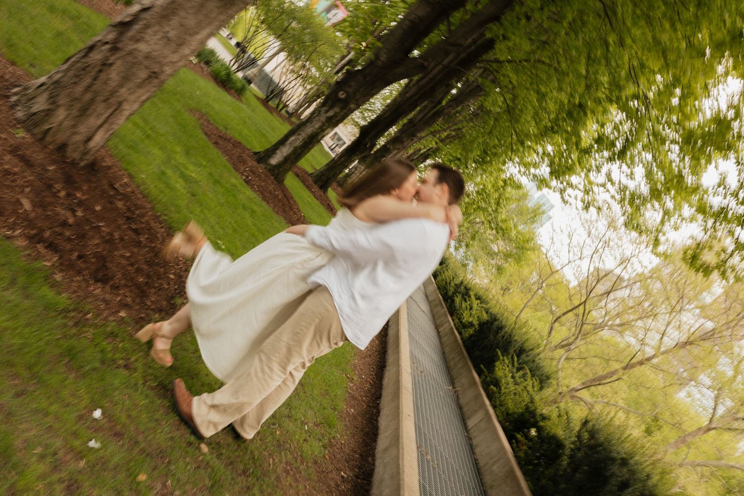 Couple sharing a romantic kiss in a lush green park, bride in white dress lifted off ground by groom.