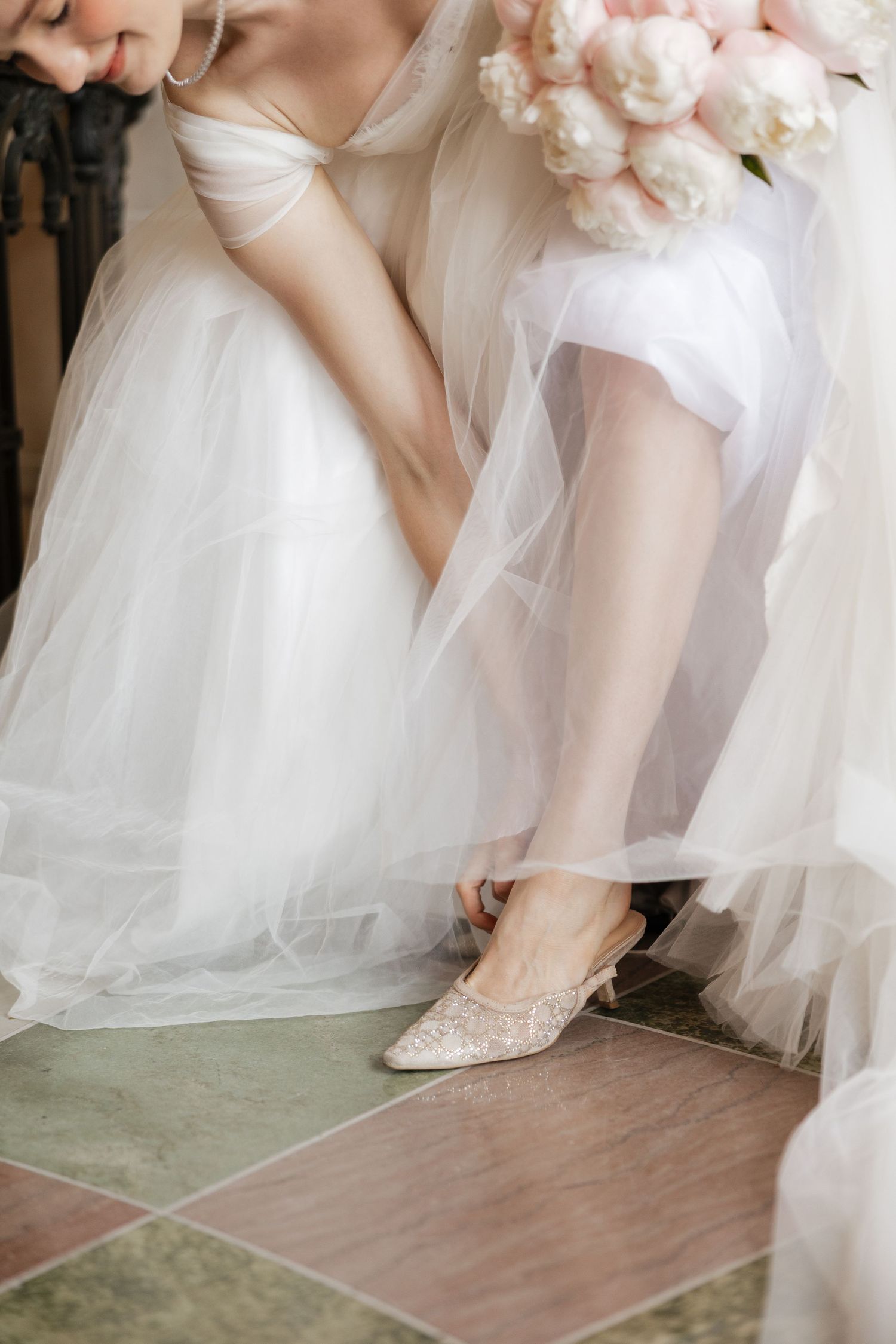 Bride in white tulle gown fastening elegant lace kitten heel shoes while holding a soft pink peony bouquet.