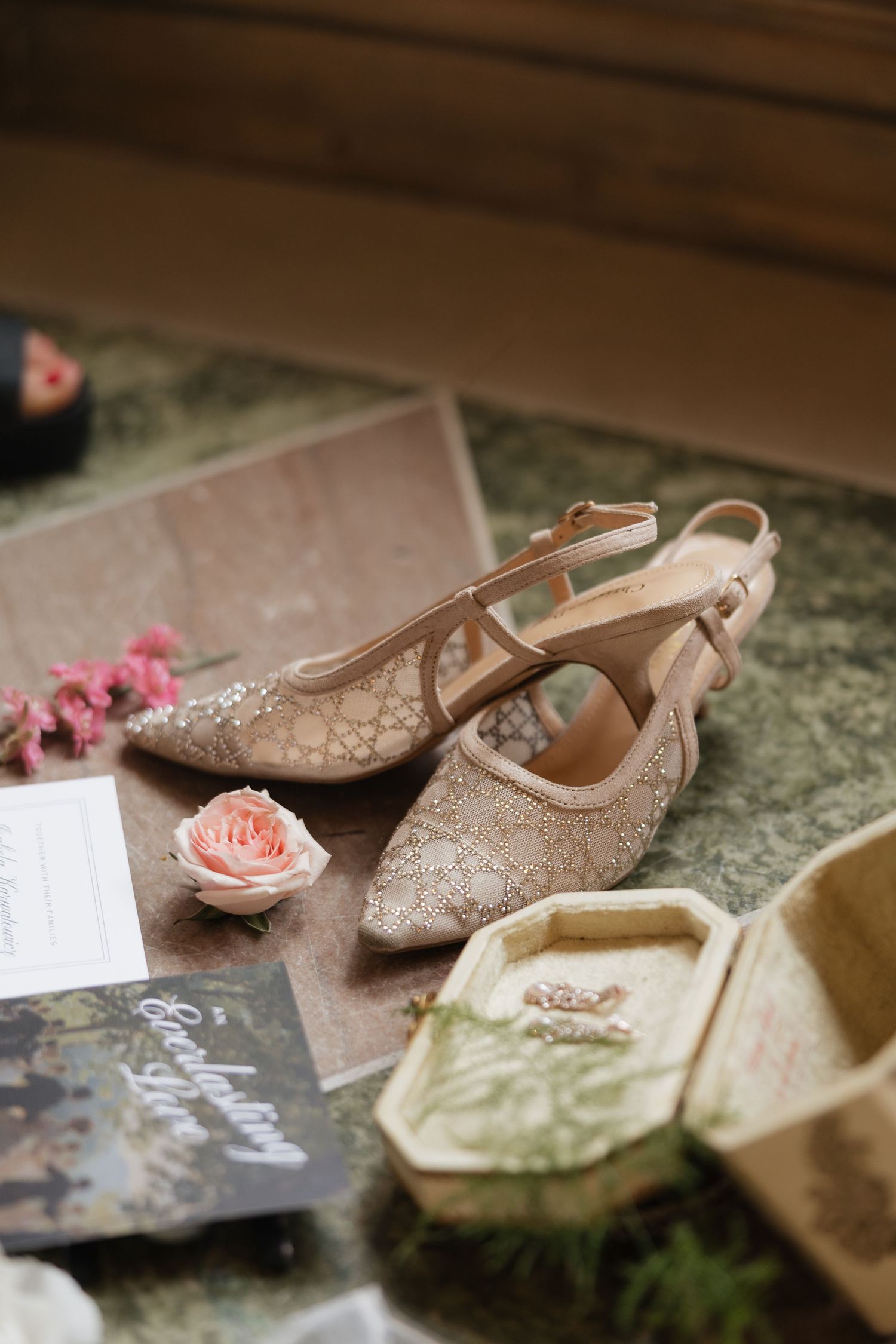 Elegant beige lace bridal heels displayed with a pink rose, wedding rings in a wooden box, and floral stationery.