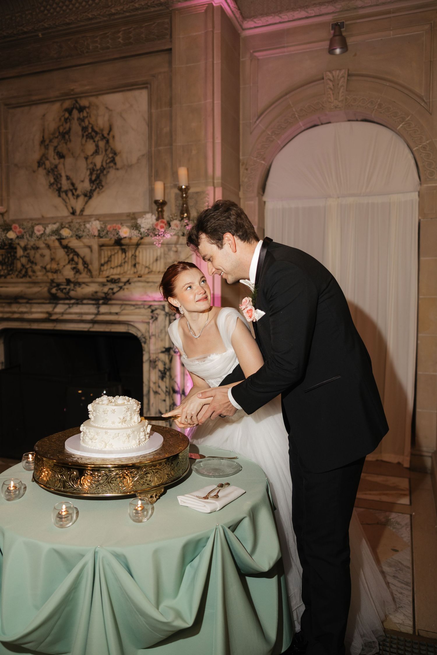 Bride and groom cutting their white wedding cake at an elegant reception venue with a marble fireplace.