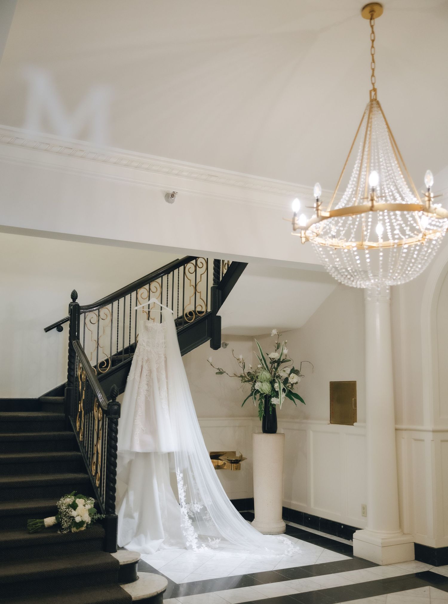 Elegant white wedding dress hangs near a grand staircase with a crystal chandelier and floral arrangements in a bright venue.