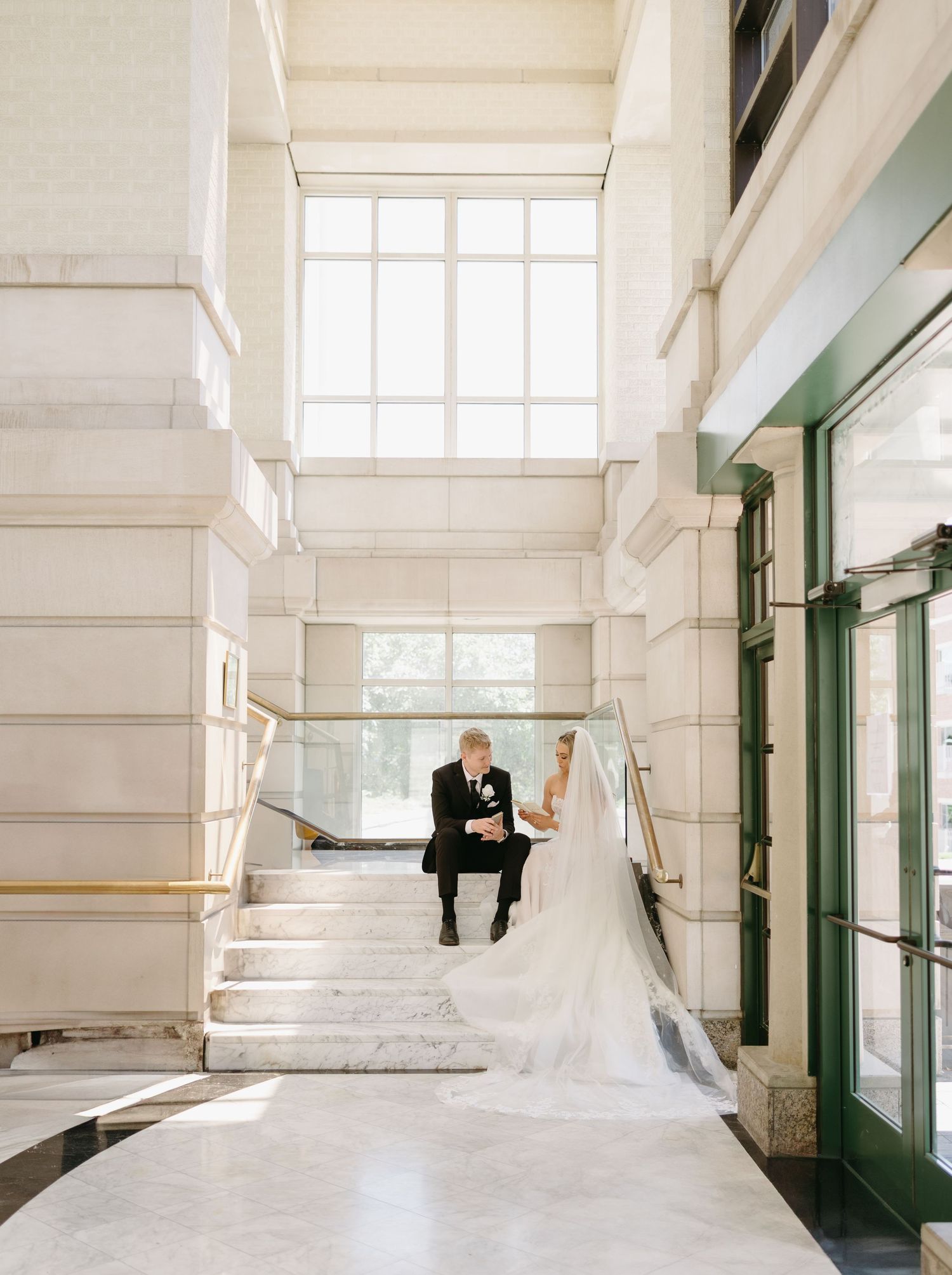 A couple sits together on white marble stairs in an elegant building interior with tall windows and green doorways.