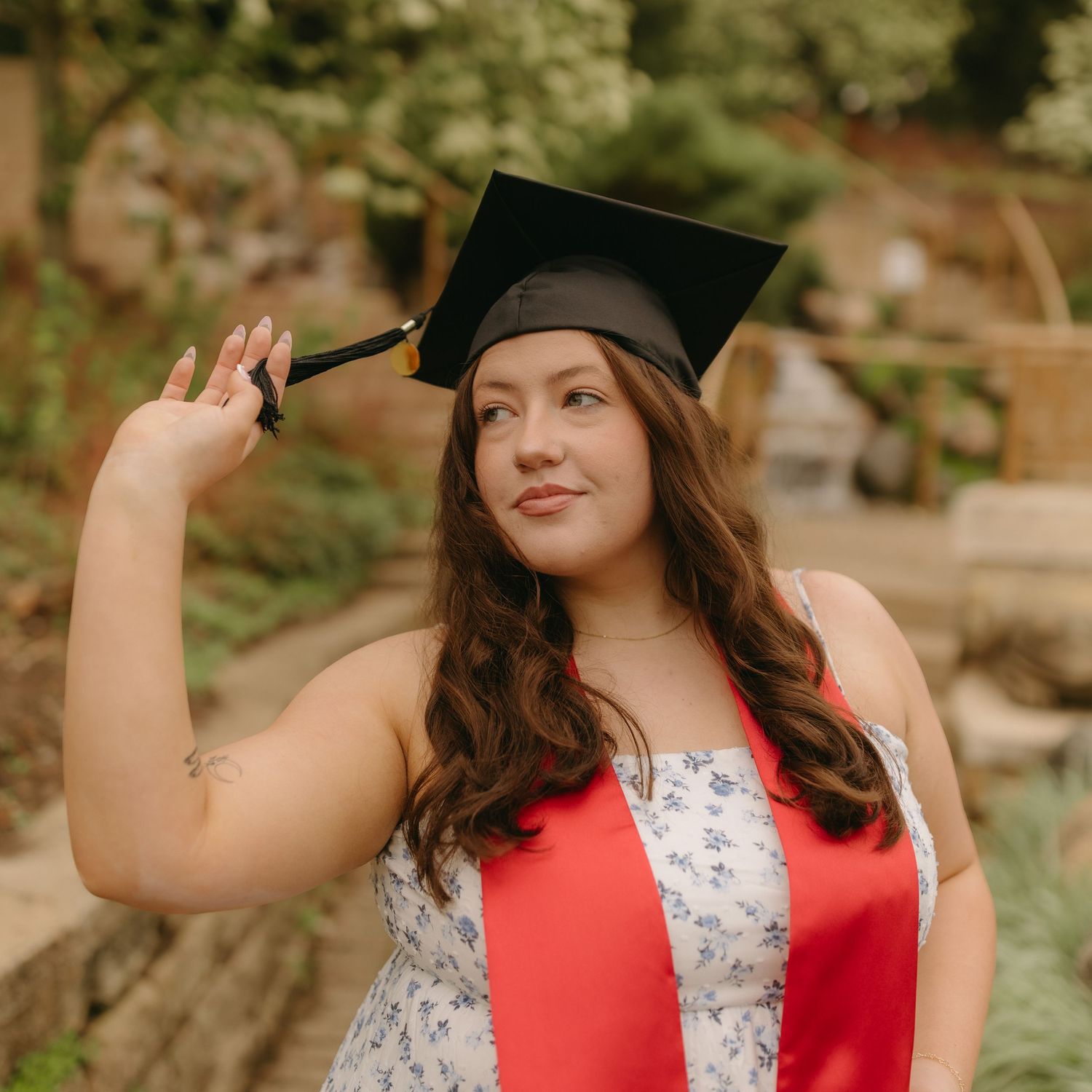 Graduate in floral dress and red stole poses outdoors with graduation cap during commencement ceremony.