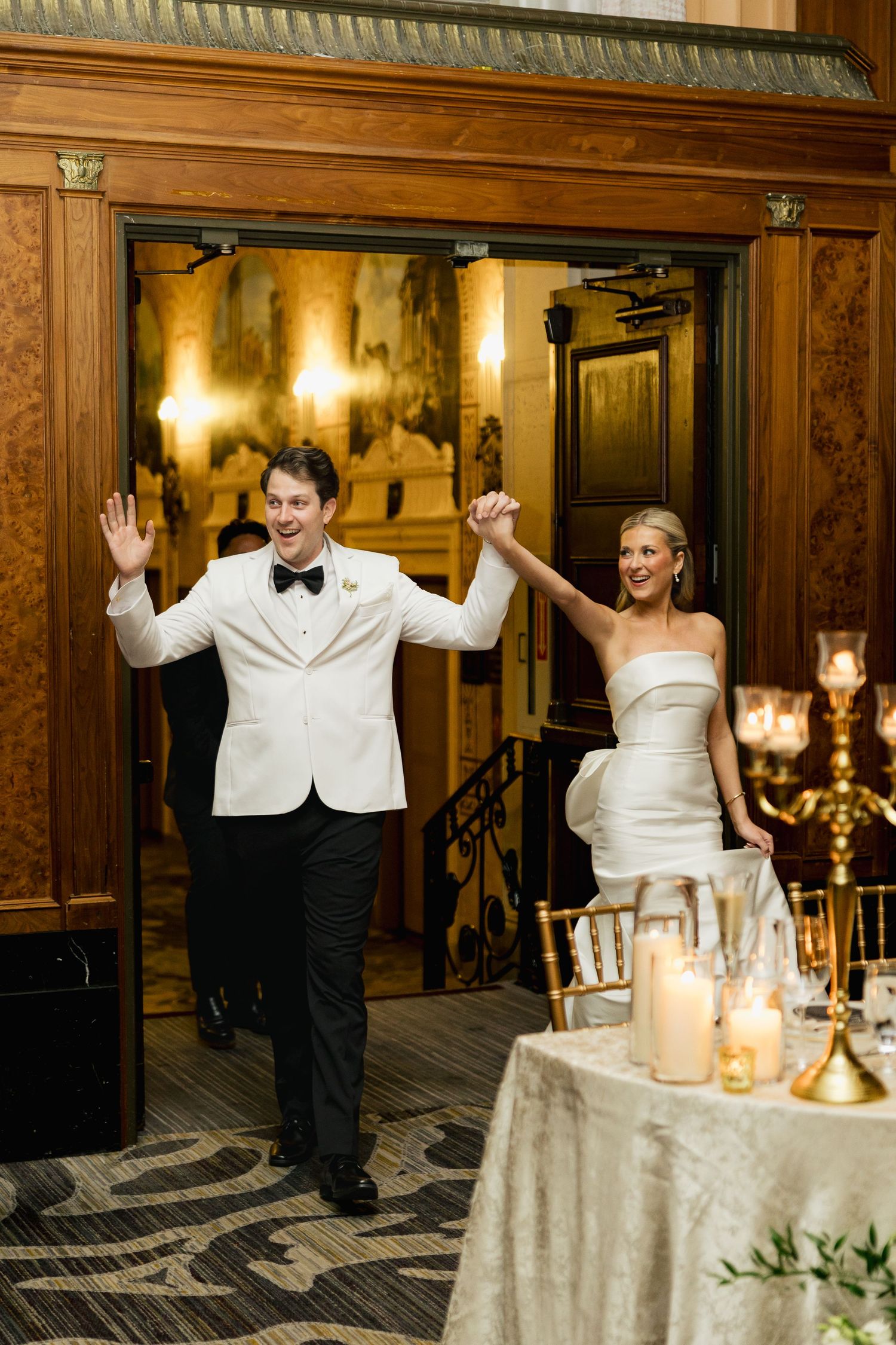 Bride and groom make grand reception entrance, groom in white tuxedo jacket, bride in strapless gown, holding hands.