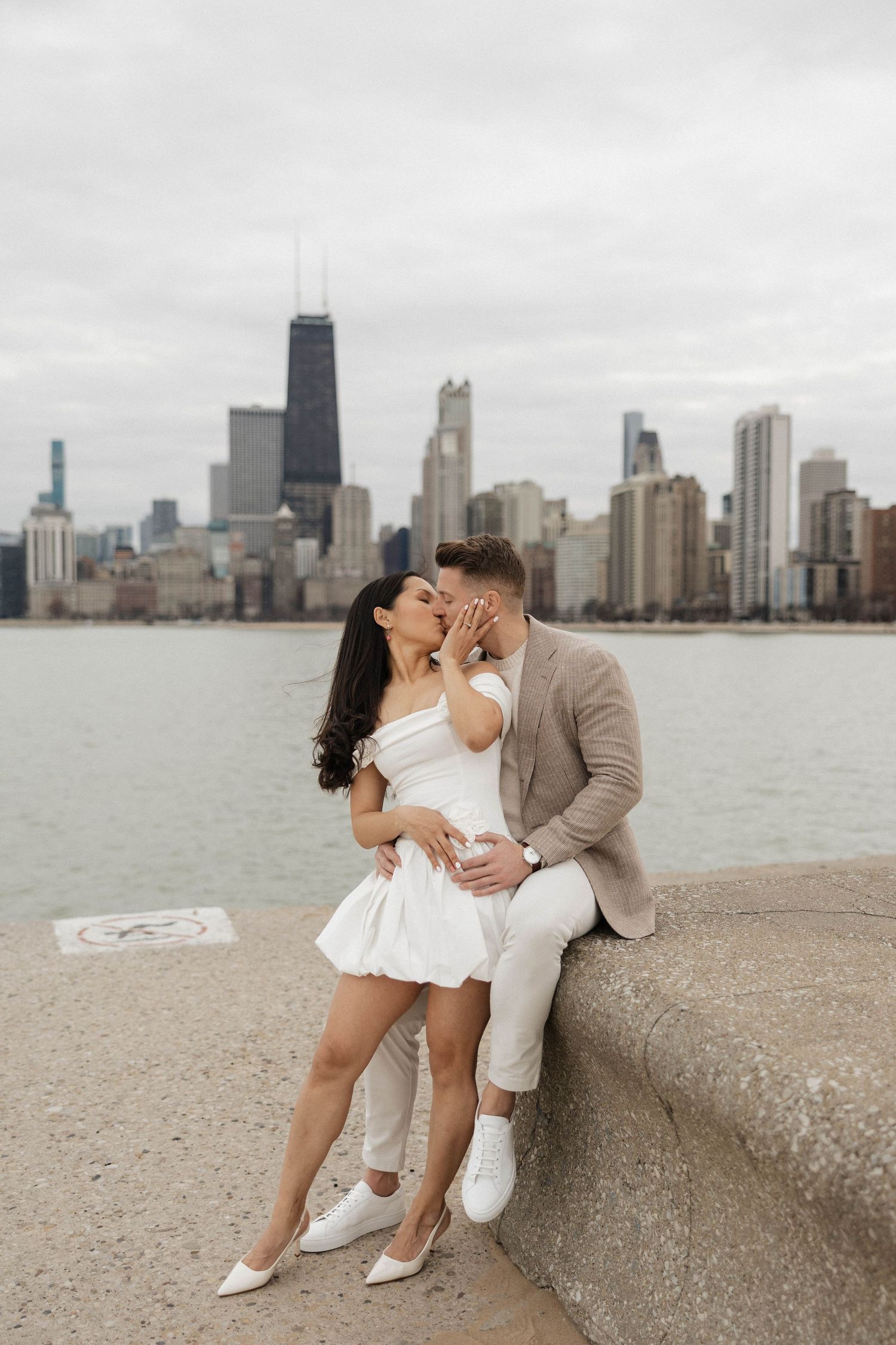 Couple kissing on Chicago lakefront seawall in white dress and tan suit with city skyline backdrop.
