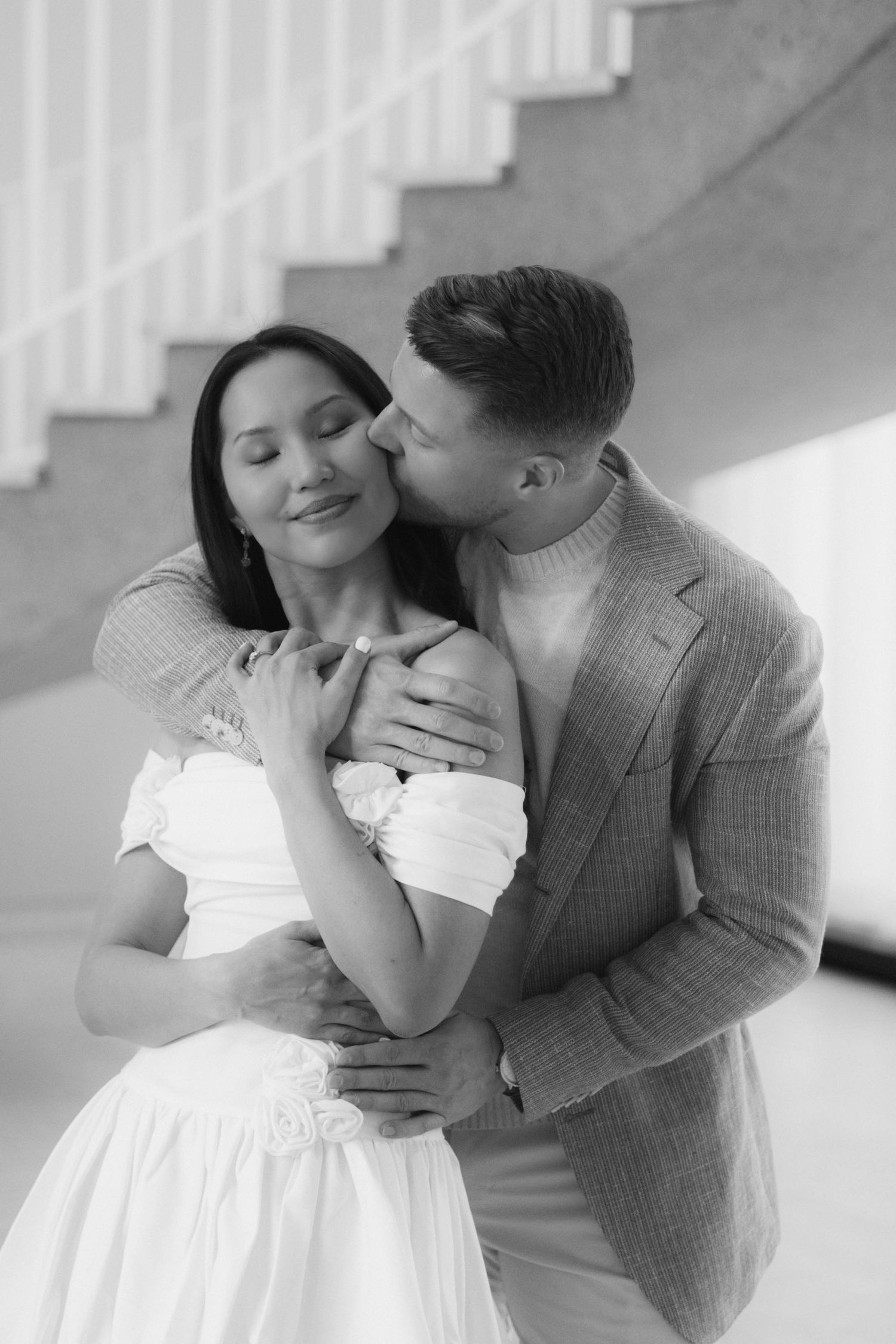 Black and white photo of a couple embracing, man kissing woman's cheek near a staircase.