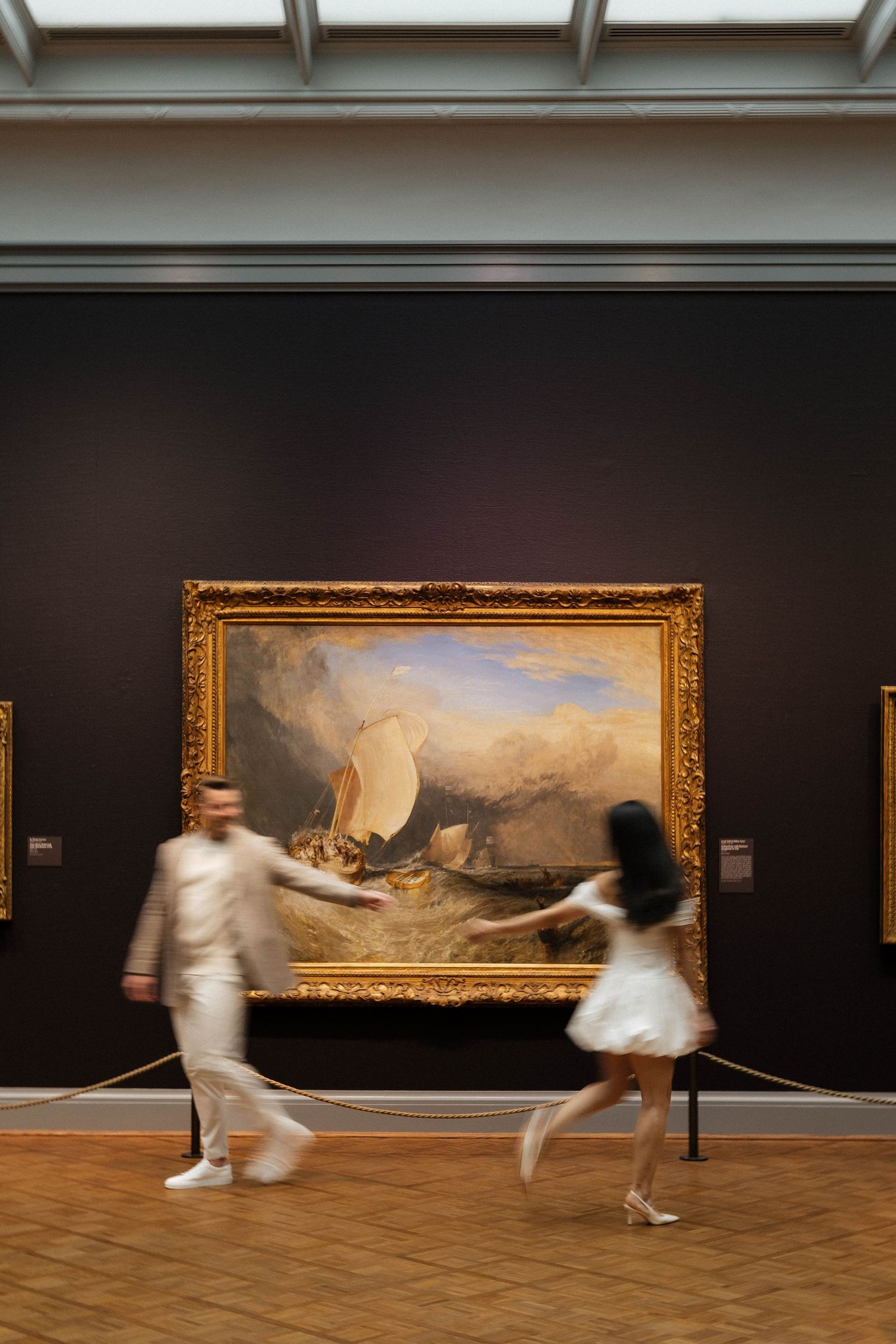 Blurred couple dancing in front of a large Turner seascape painting in a museum gallery.