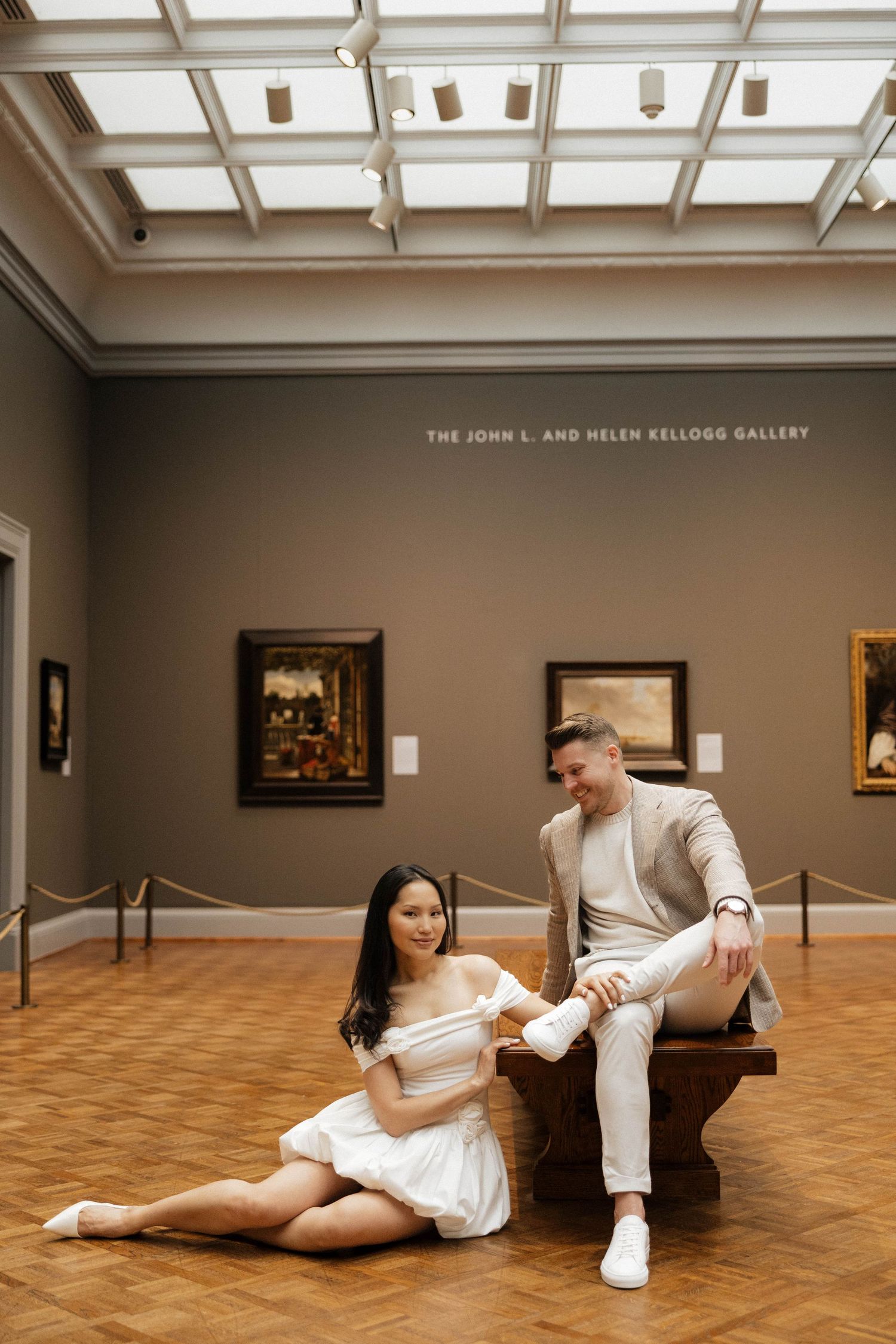 Couple poses in the John L. and Helen Kellogg Gallery, woman in white dress sits on floor beside man on bench.