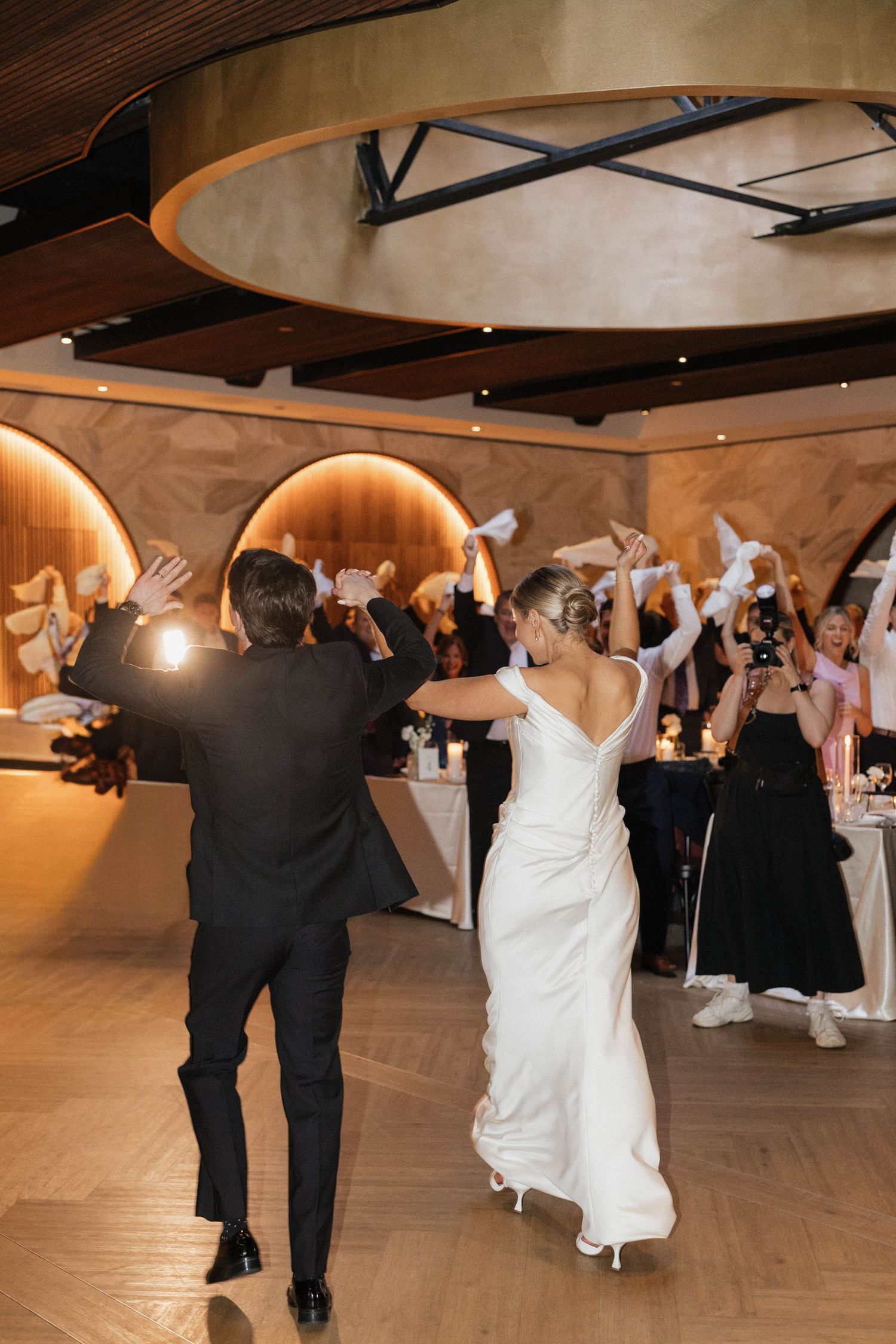 Bride and groom make grand entrance at elegant wedding reception as guests wave white napkins in celebration.