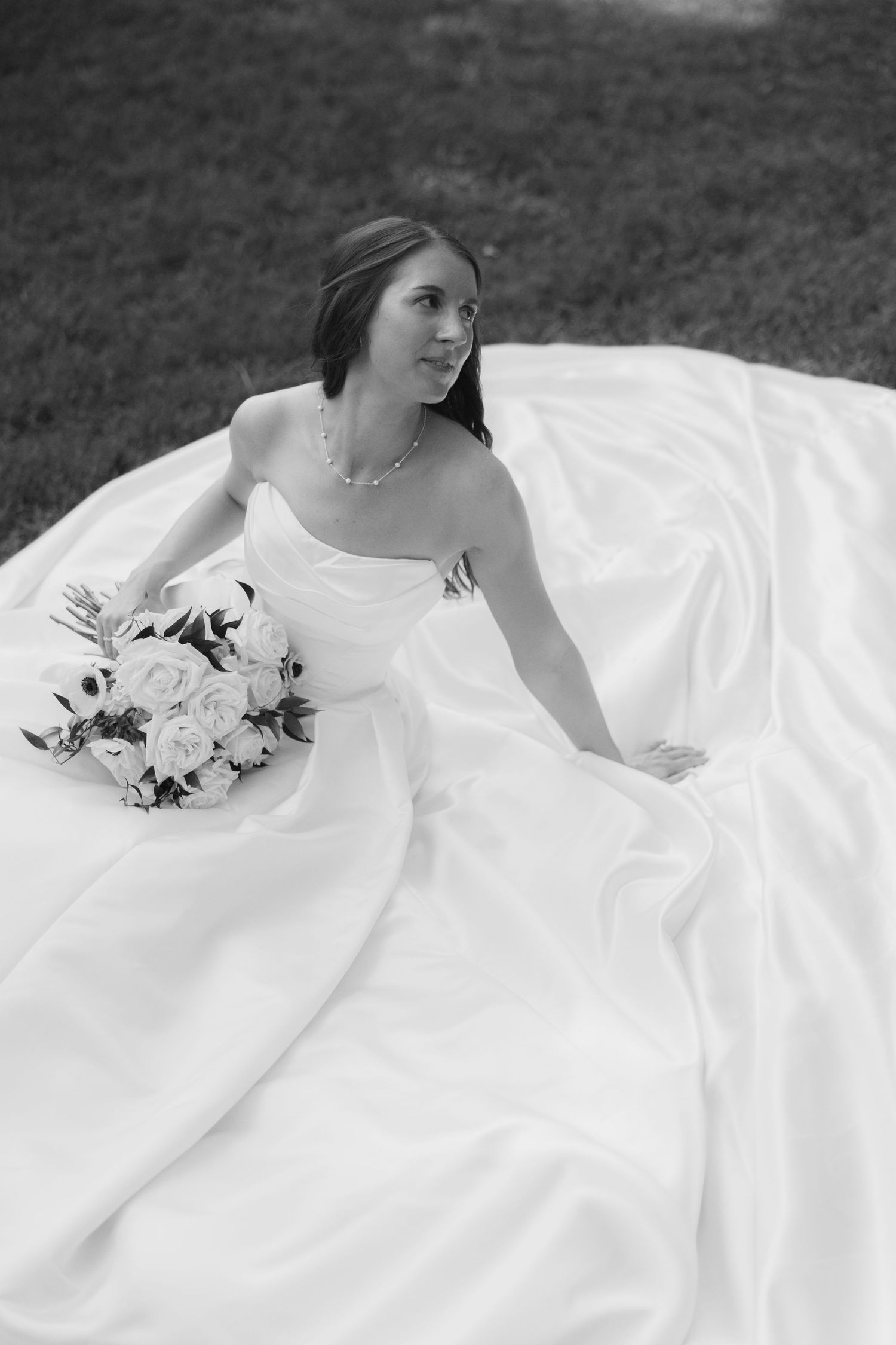 Black and white photo of bride in strapless ball gown sitting on grass with white rose bouquet outdoors.
