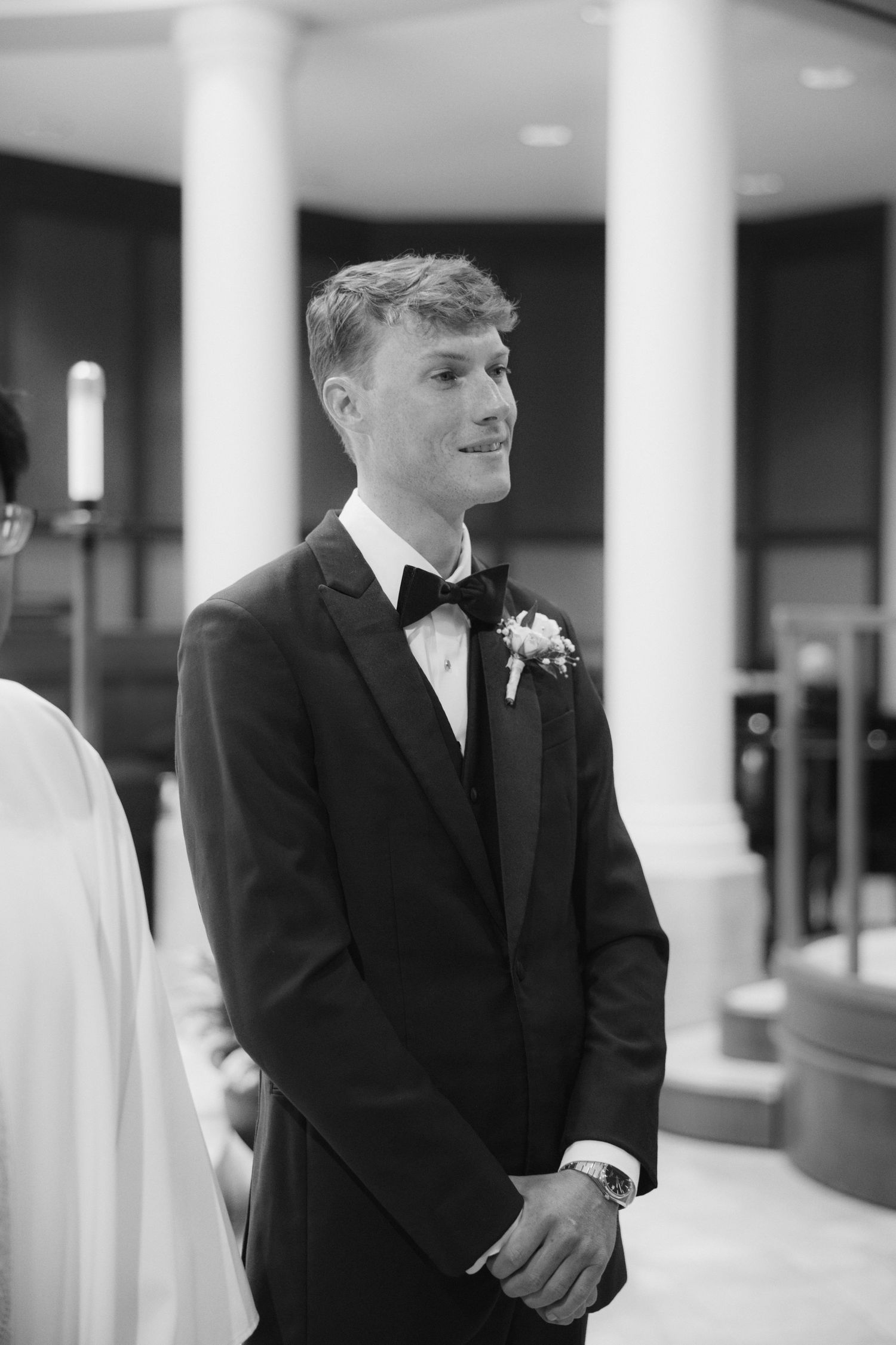 Groom in black tuxedo with boutonniere smiling at altar during wedding ceremony, black and white photo.