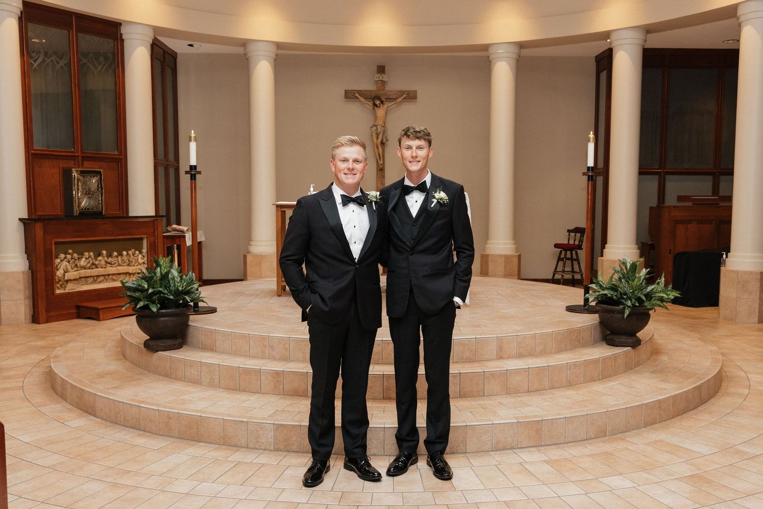Two men in tuxedos posing in a Catholic church with altar and crucifix backdrop.