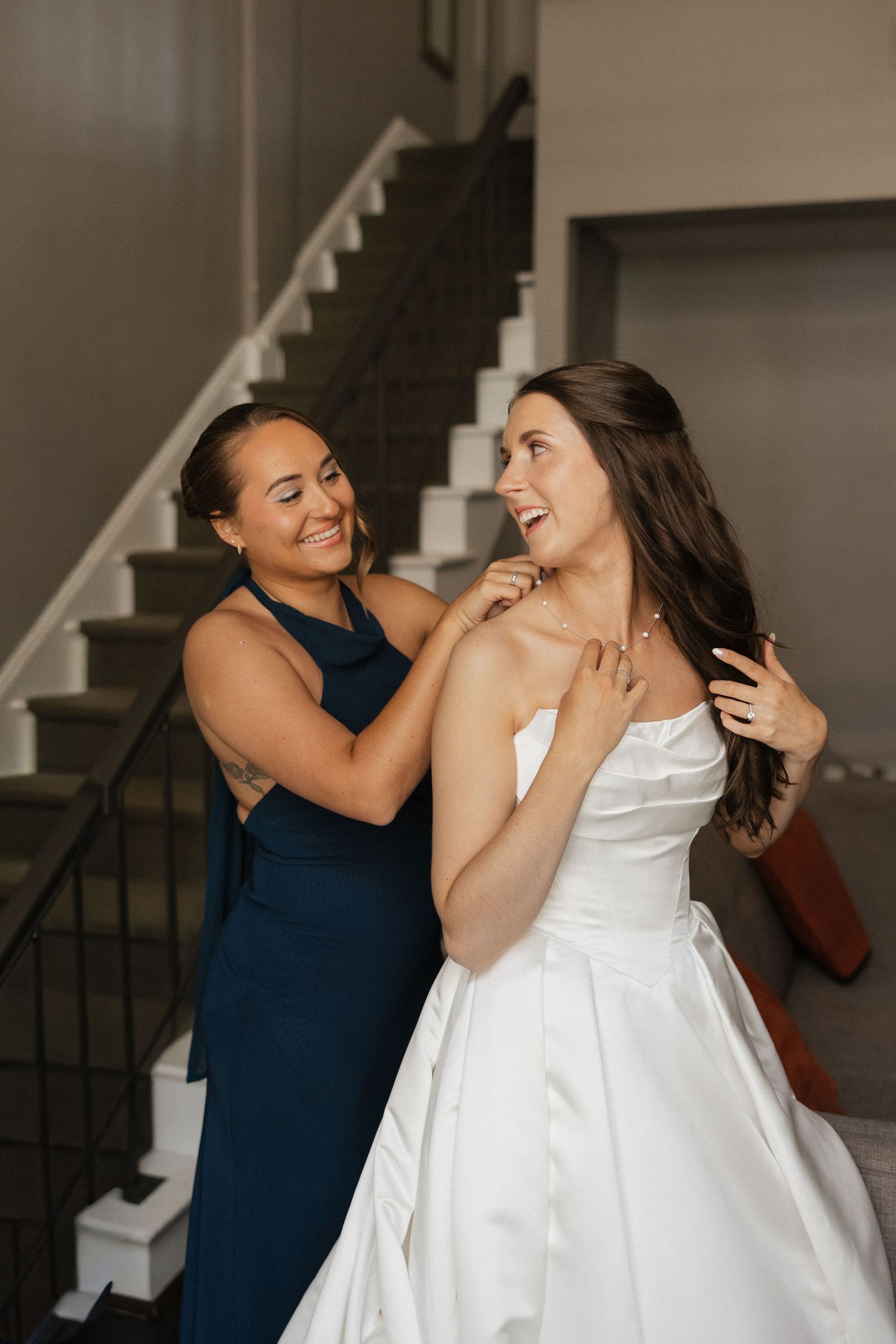 A bridesmaid in a navy dress helps a smiling bride in a white gown with her necklace near a staircase.