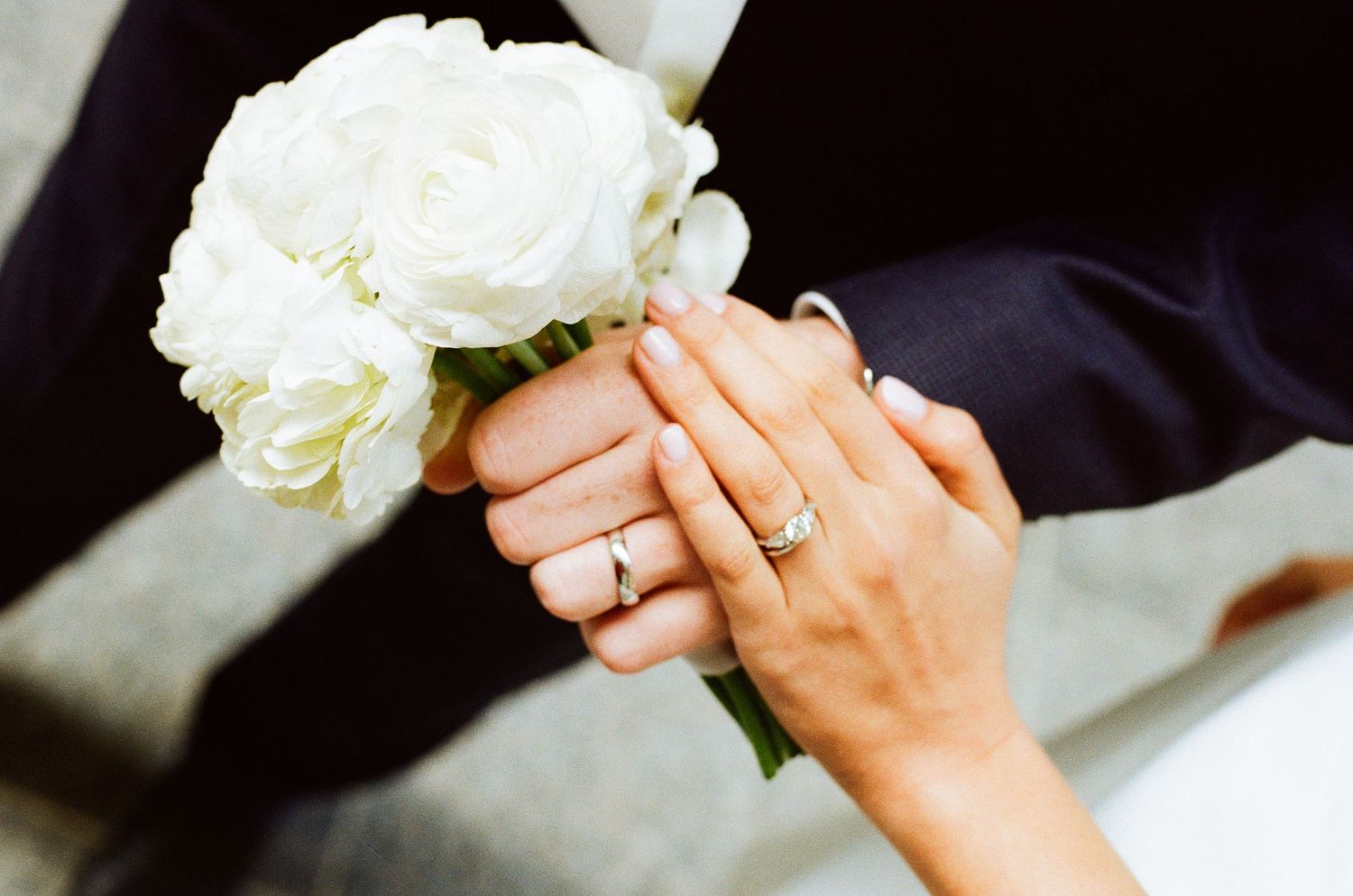 Bride and groom holding white rose bouquet with wedding rings visible on their hands.