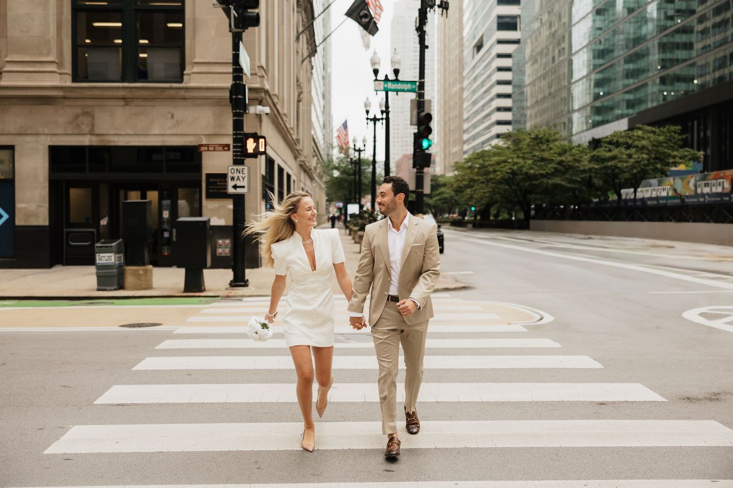 Happy couple holding hands while crossing a city street crosswalk, woman in white dress, man in tan suit.