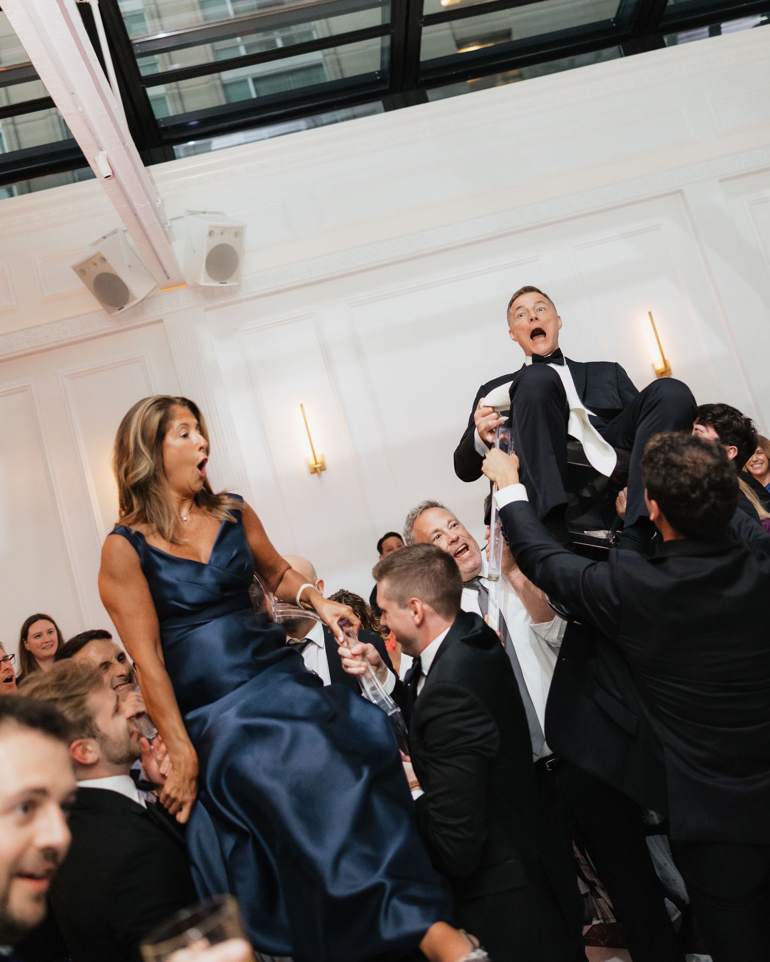 Guests lift bride in blue gown and groom in tuxedo during energetic hora dance at wedding reception.