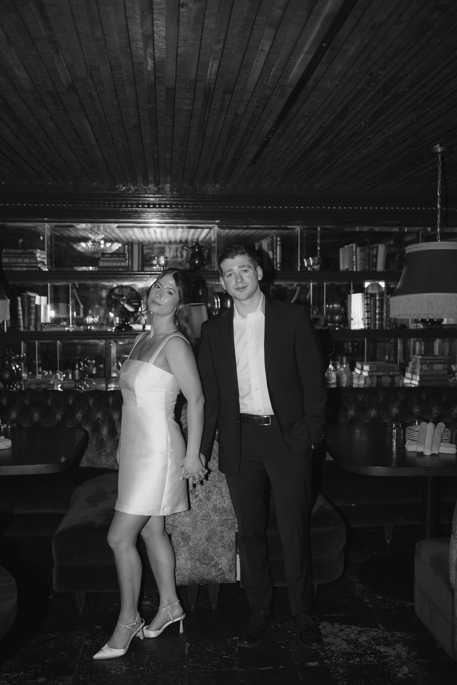 A couple in formal attire stands together in a dimly lit venue with wood paneled ceiling.