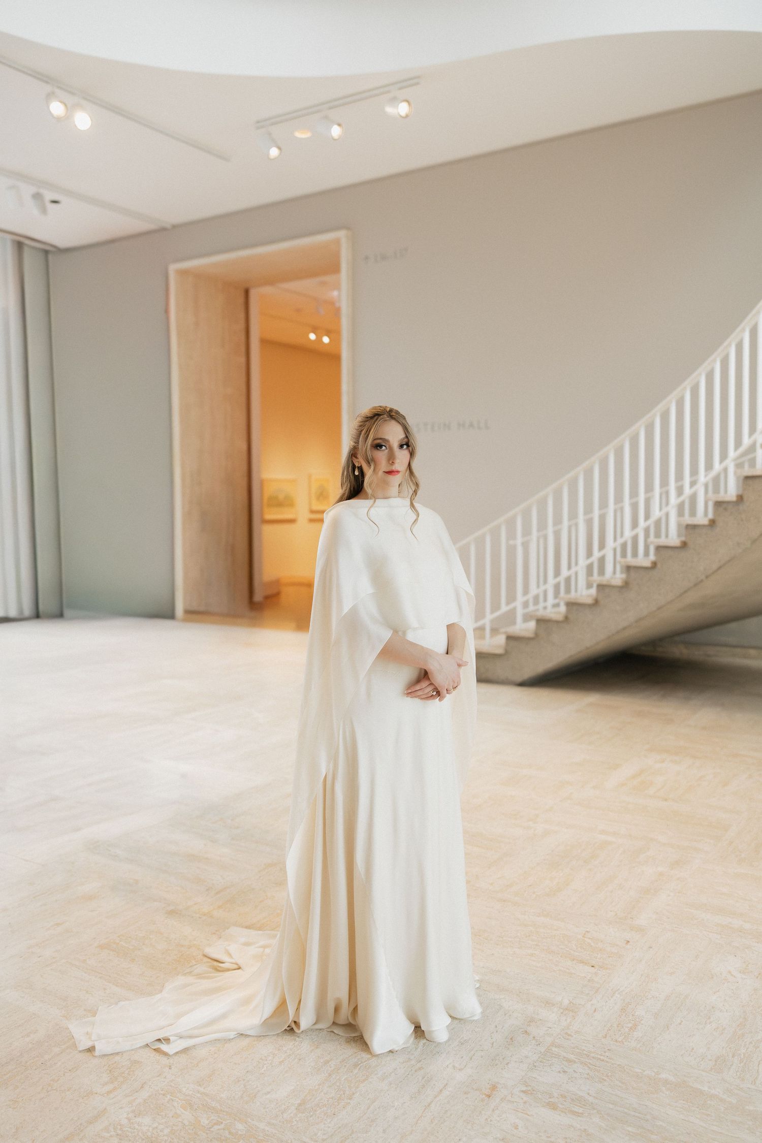 A bride in an elegant white long-sleeved wedding gown stands gracefully in front of a staircase with dramatic lighting.