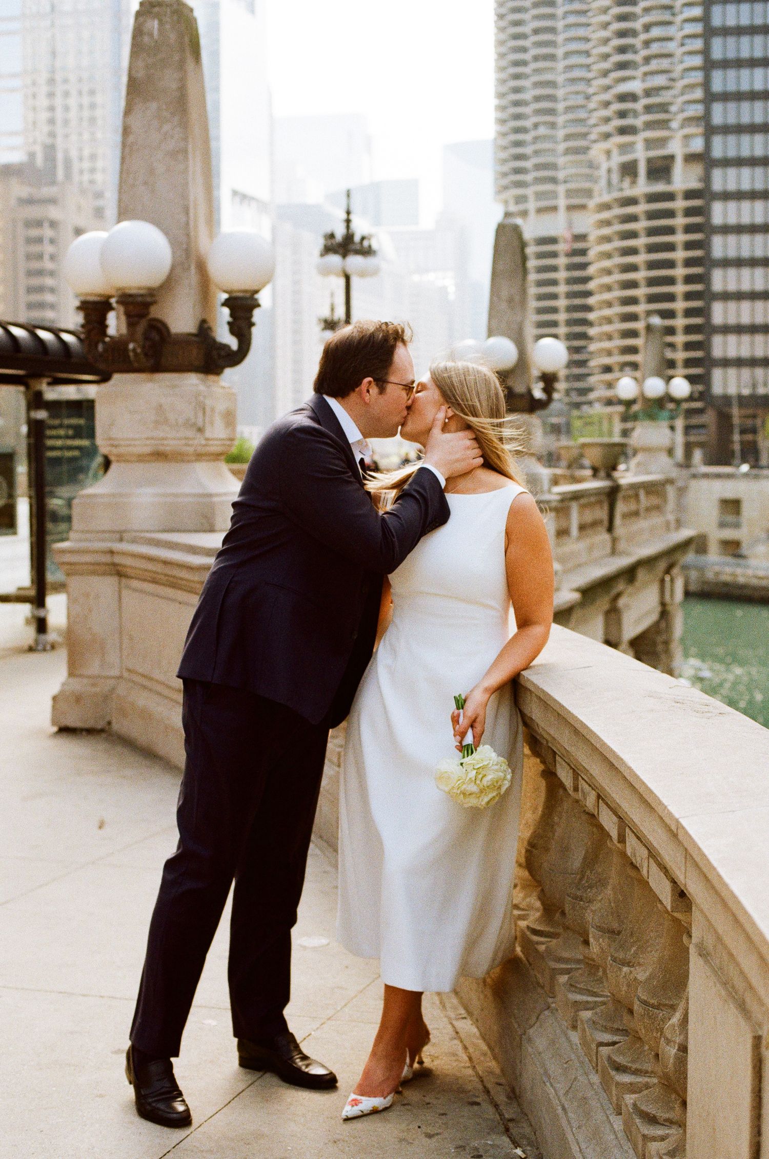 Bride and groom share a kiss on a stone bridge overlooking the Chicago River, bride holding white bouquet in midi dress.