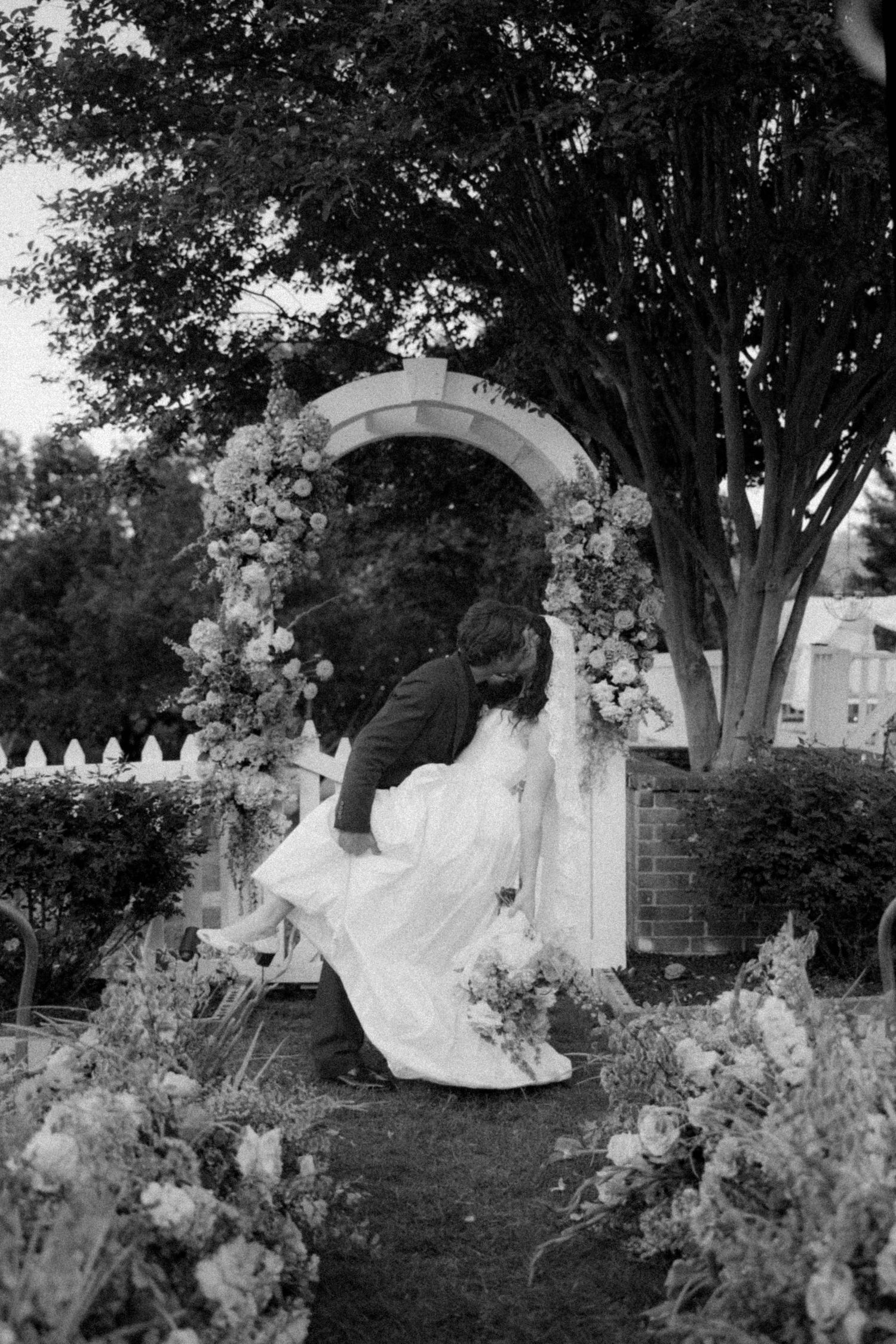 Black and white photo of a couple sharing a romantic dip kiss under a floral arch in a garden wedding.