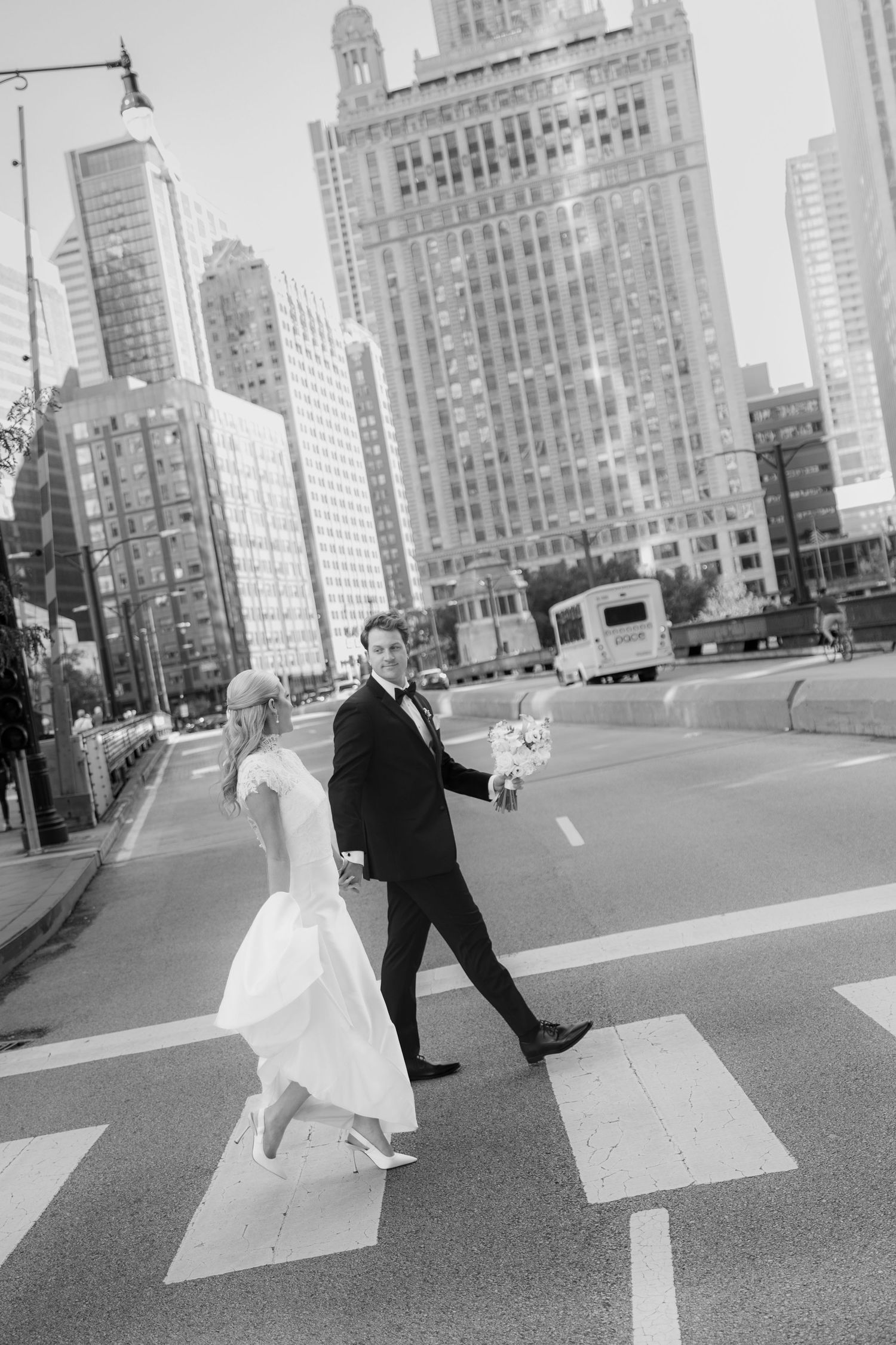 Black and white photo of a bride and groom holding hands and crossing a city crosswalk with tall skyscrapers behind them.