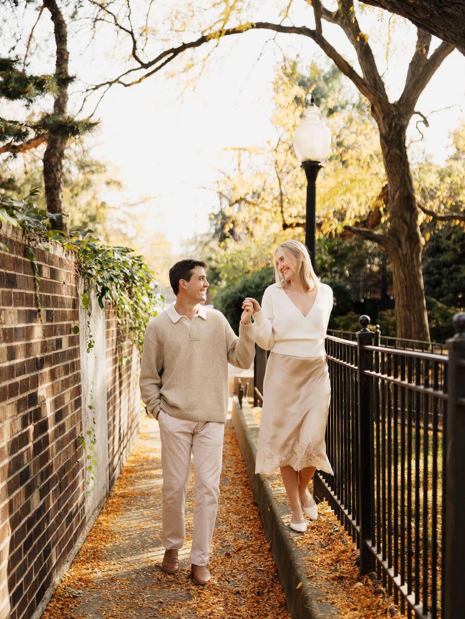 Smiling couple in neutral outfits holds hands while walking along a leaf-covered brick path beside an iron fence in warm autumn light.