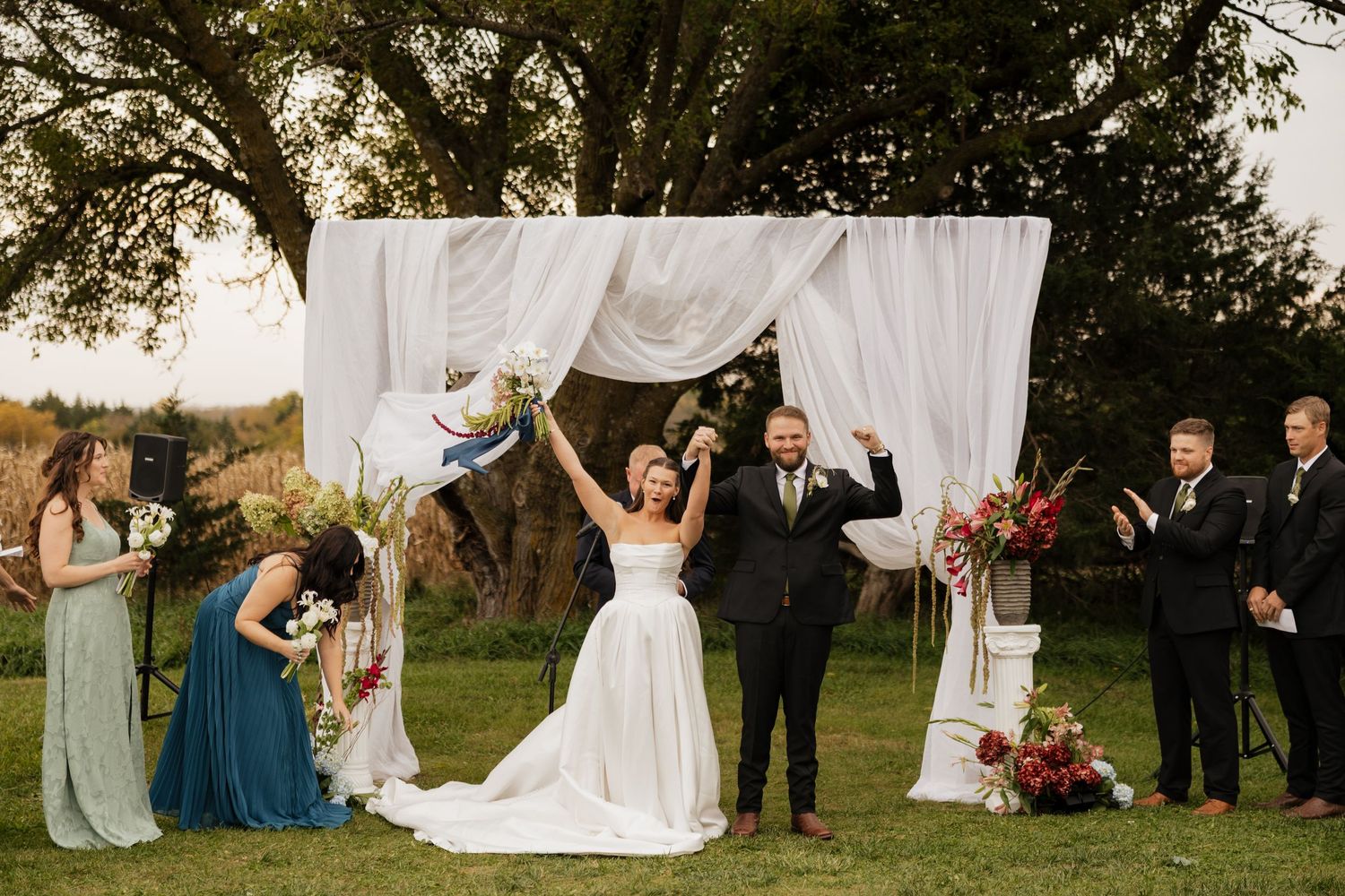 Newlywed bride raises her arms in celebration beside her groom under a draped ceremony arch as bridesmaids and groomsmen cheer.