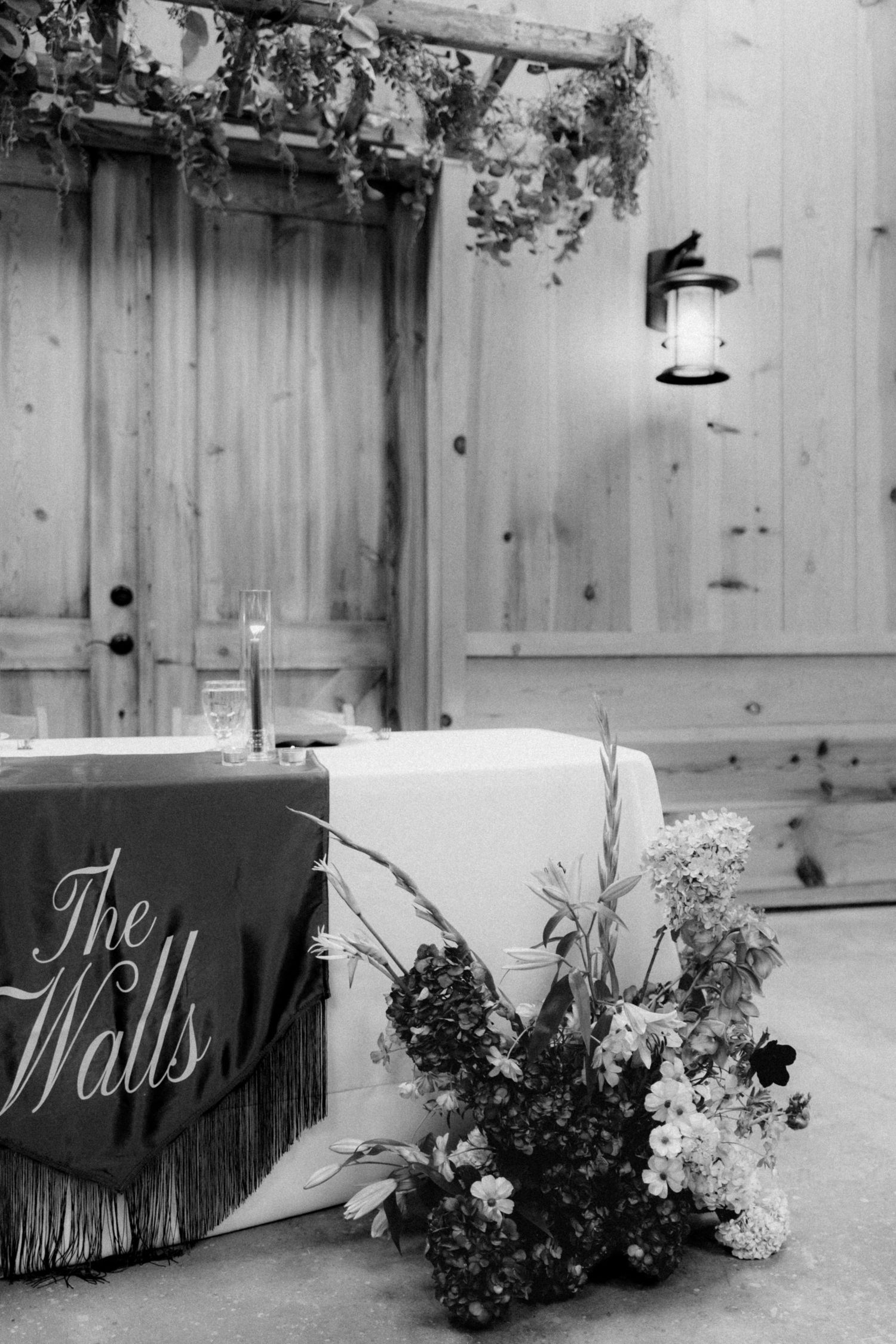 Black and white rustic barn wedding sweetheart table with a draped banner reading The Walls, floral arrangements, and a hanging lantern.