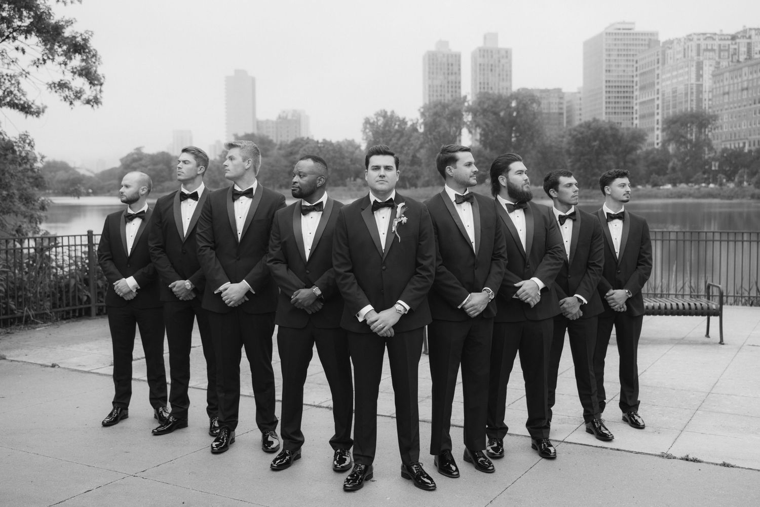 Groom and groomsmen in black tuxedos and bow ties pose outdoors with Chicago skyline in black and white wedding photo.