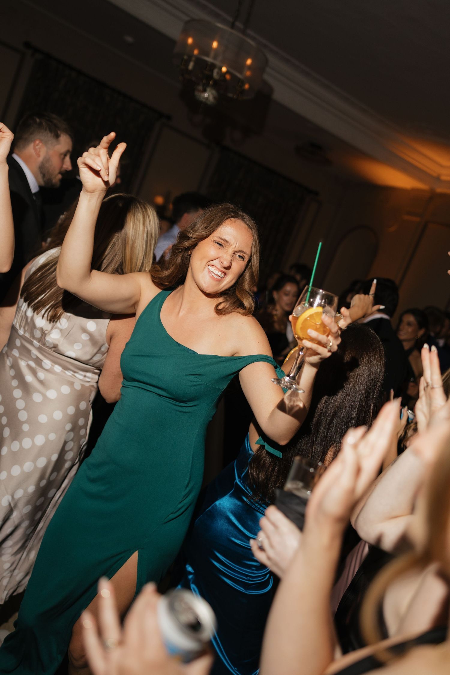 Woman in green off-shoulder dress dancing joyfully at elegant reception, holding cocktail with raised arms.