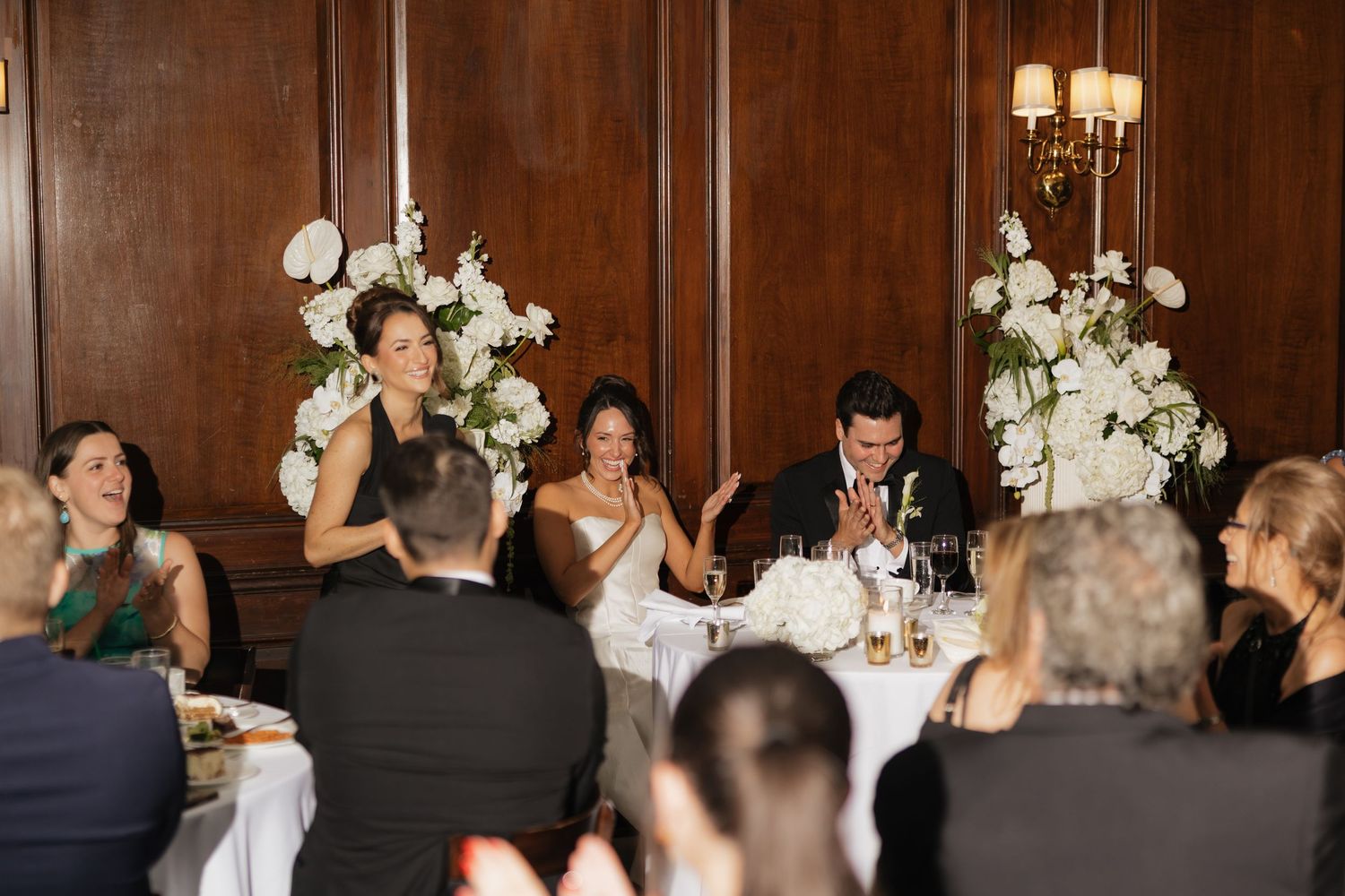 Wedding reception head table with bride and groom, white floral arrangements, and guests applauding in elegant wood-paneled venue.