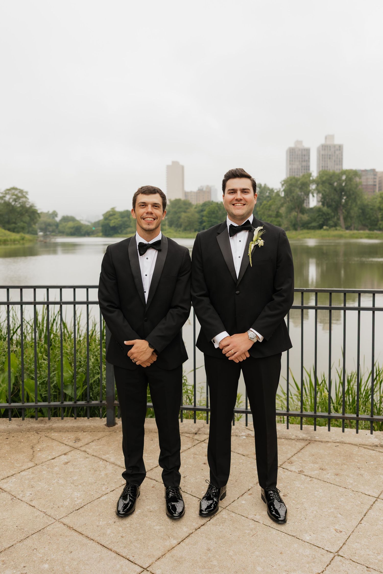 Two groomsmen in black tuxedos with bow ties pose by a lake in a park with city buildings in the background.
