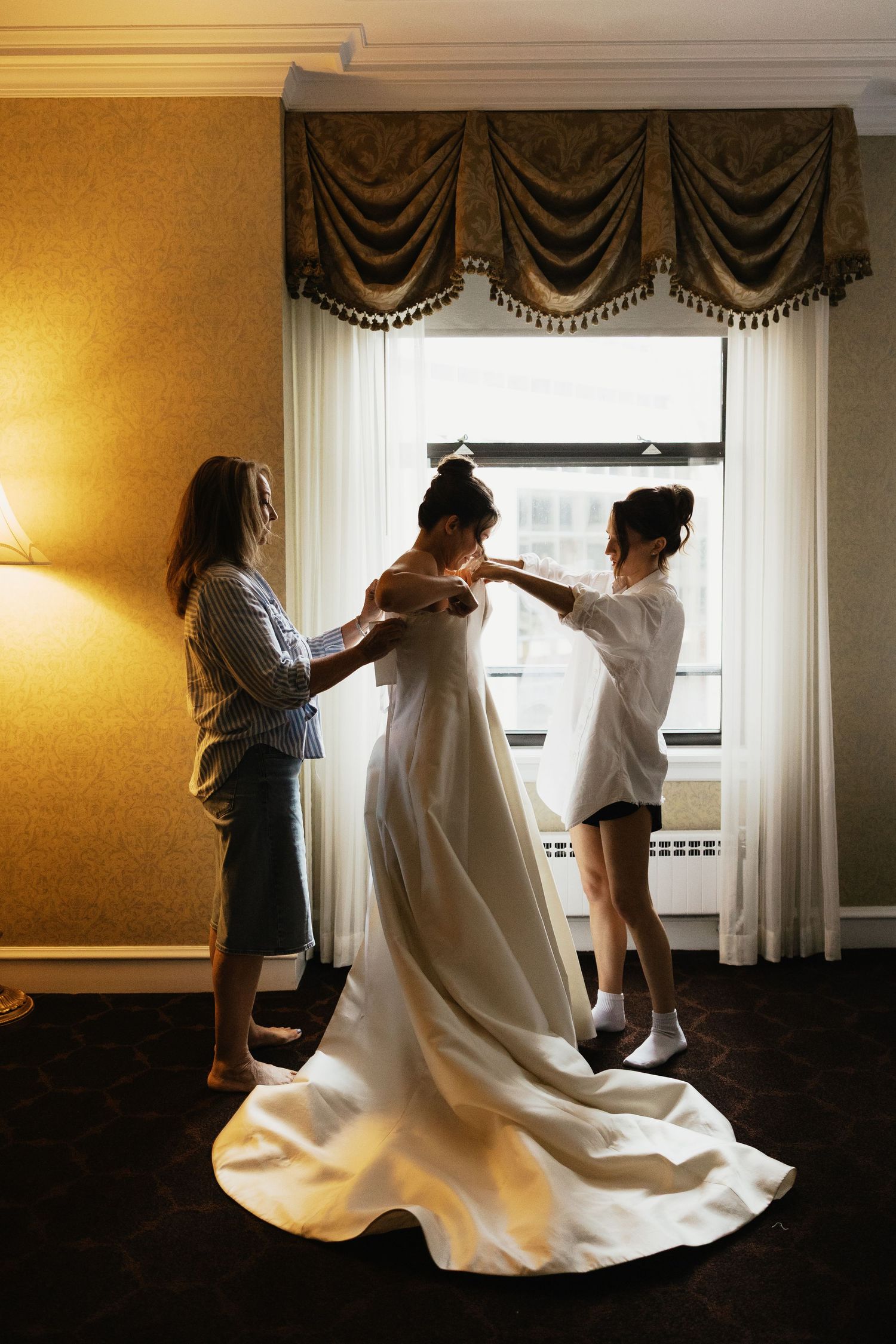 Bride being helped into her white wedding gown by two bridesmaids in a warmly lit hotel room.