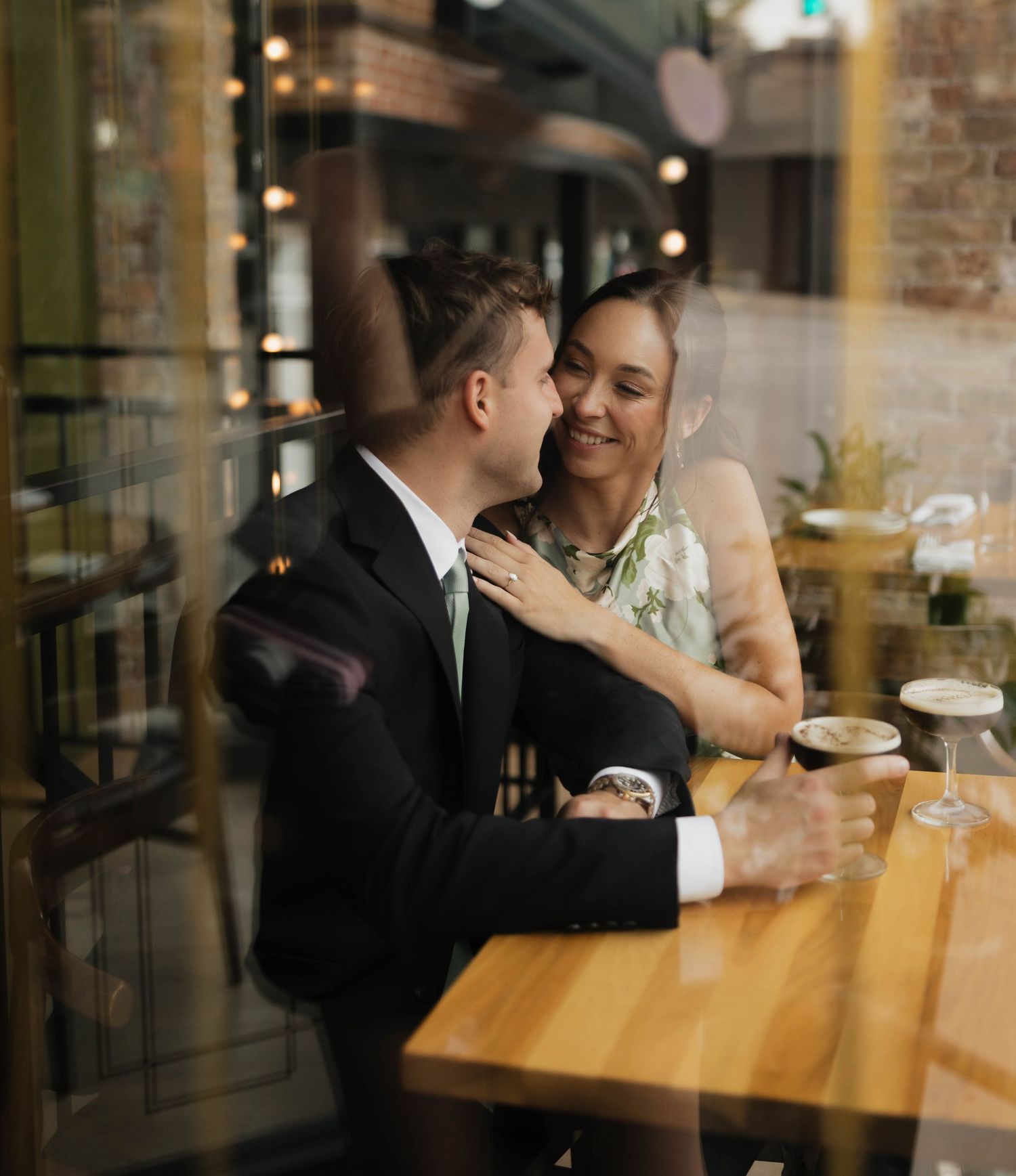 A couple shares a romantic moment at a wooden cafe table, the man in a suit kissing the smiling woman's cheek.