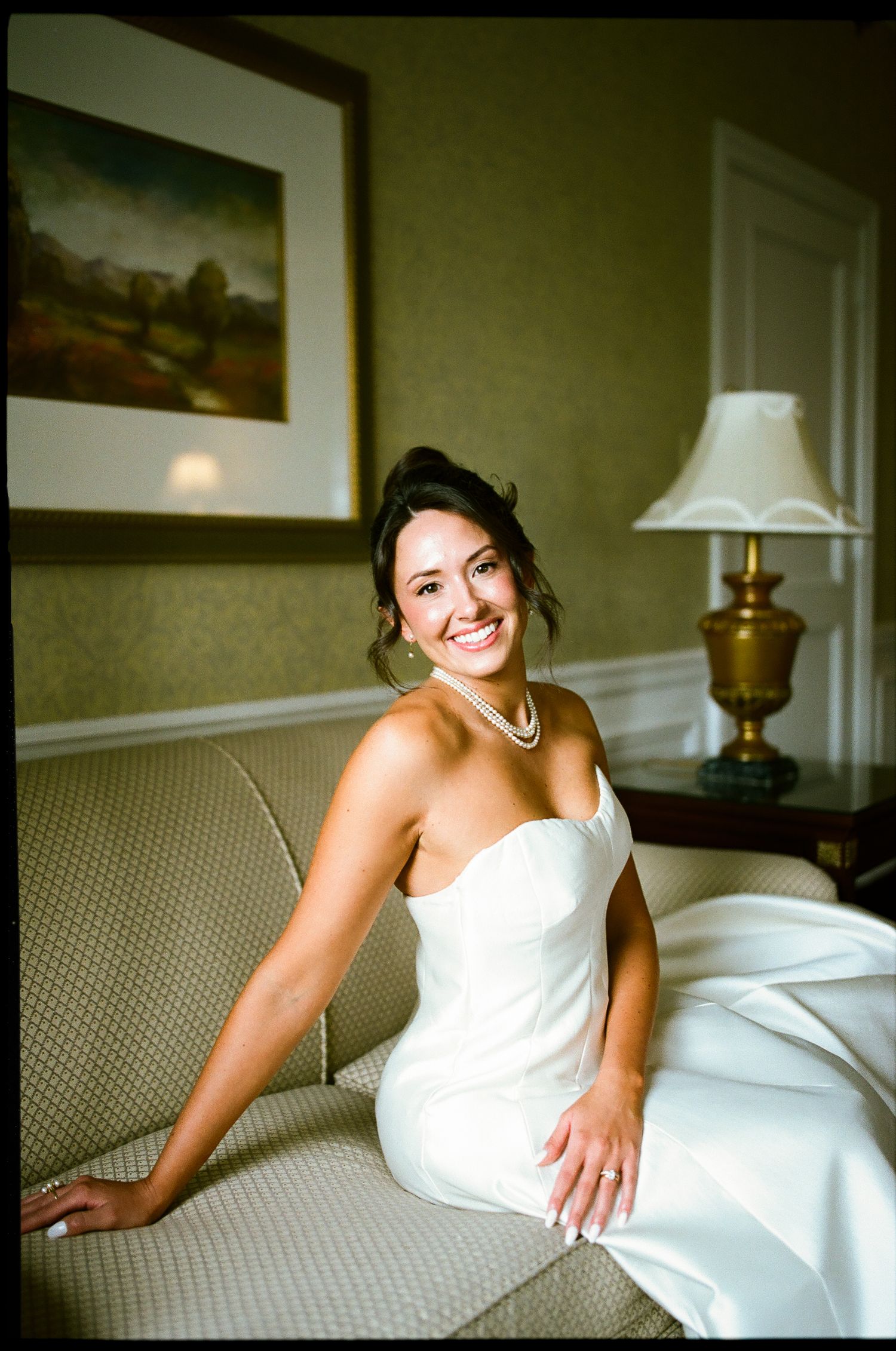 A radiant bride in a strapless white gown and pearl necklace smiles while seated on an elegant sofa.
