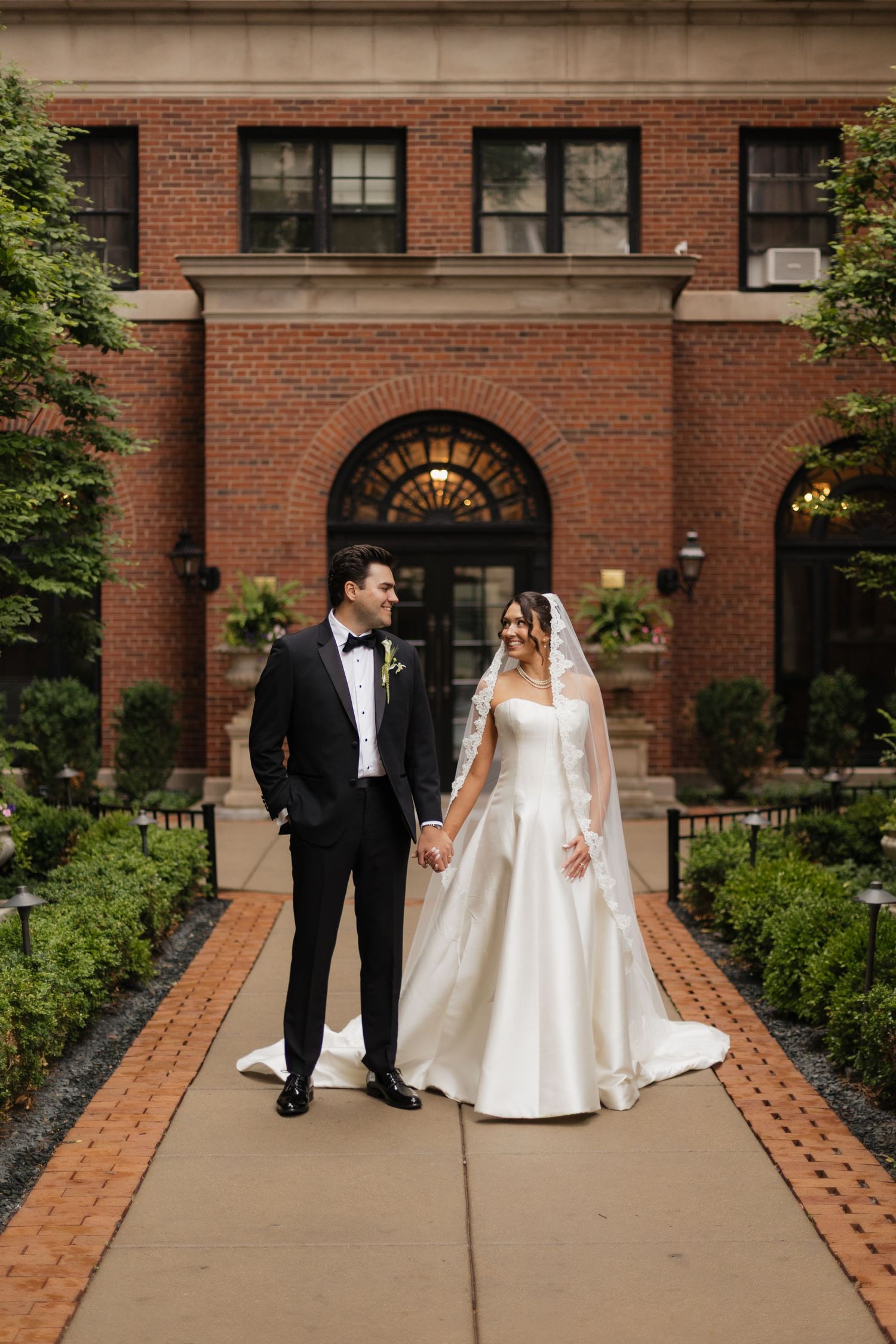 Bride and groom holding hands on a brick pathway outside an elegant red brick building on their wedding day.