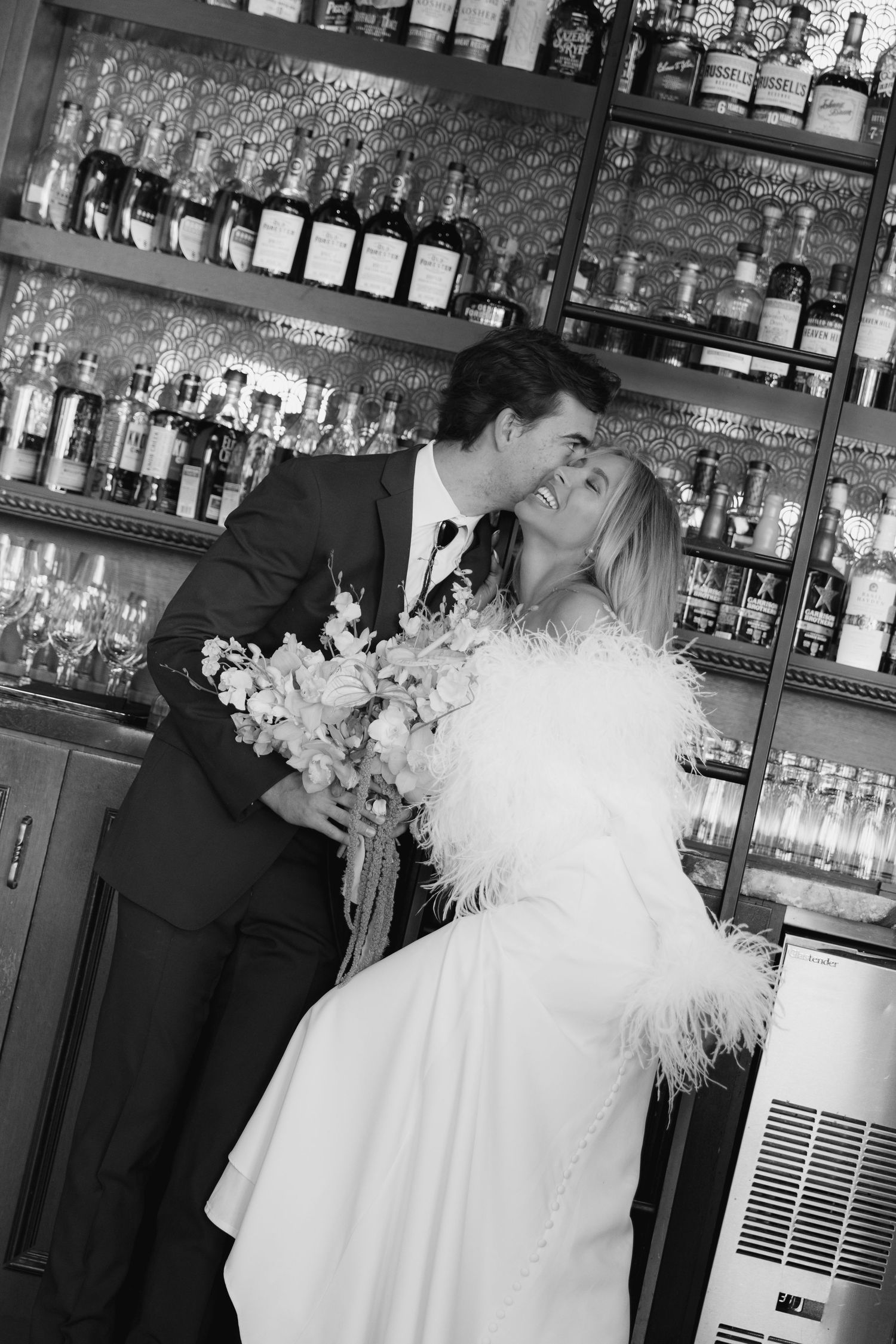 A romantic black and white photo of a couple sharing an intimate moment at a bar with bottles in the background.