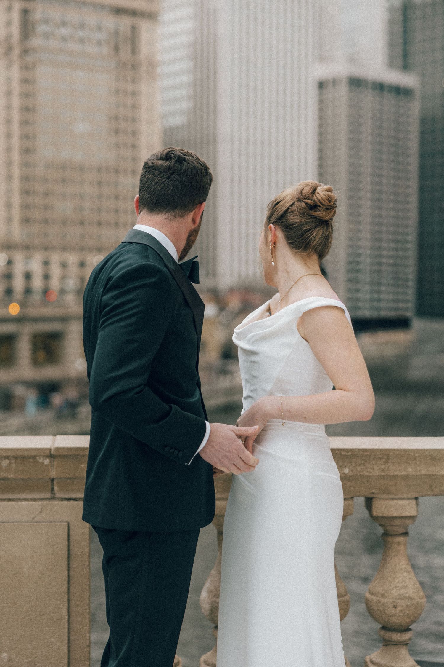 A bride and groom share a romantic moment along a stone balustrade in an urban setting with city buildings in background.
