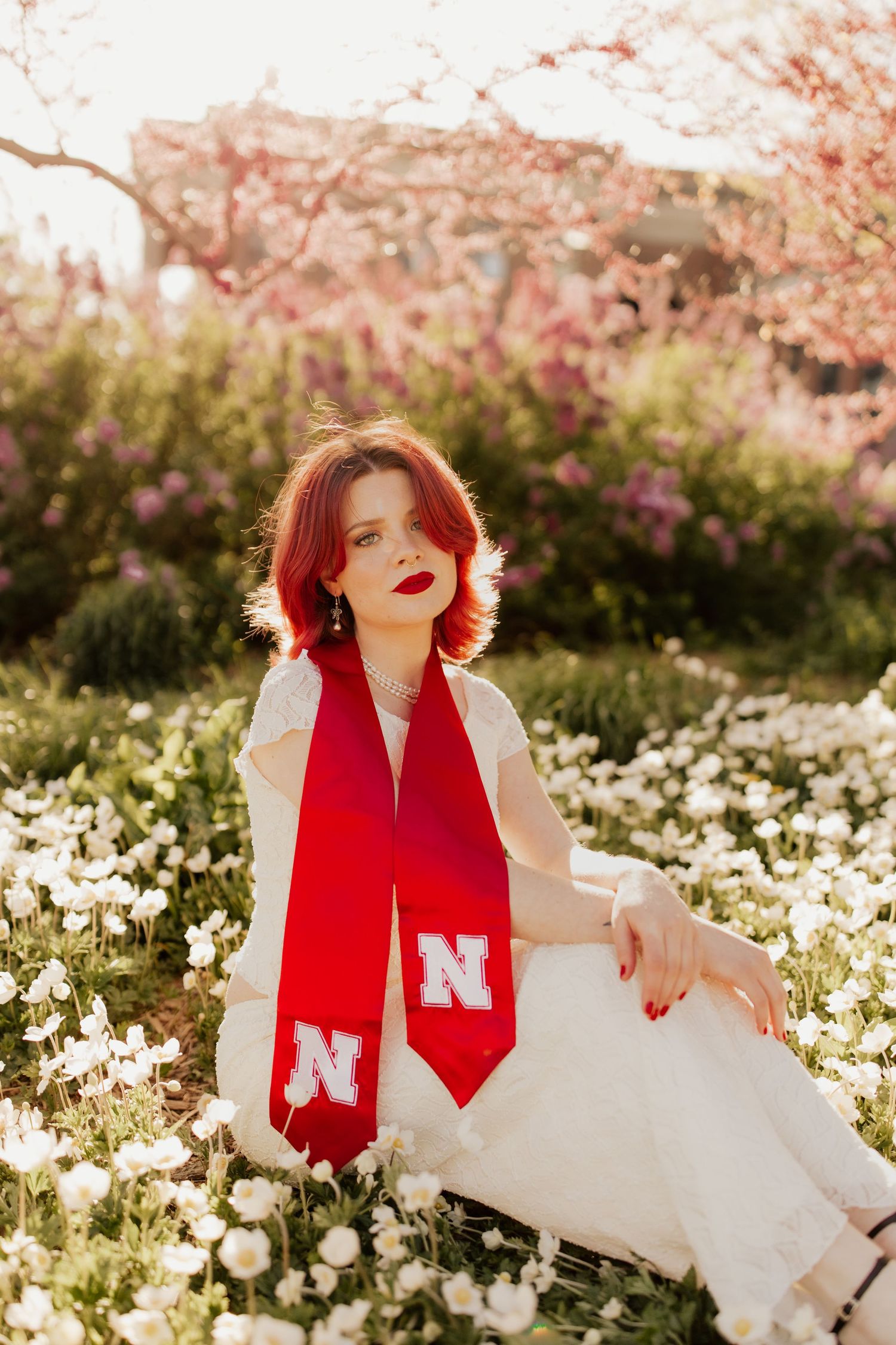 A person in a white dress and red Nebraska graduation stole sits in a blooming spring meadow of white flowers.