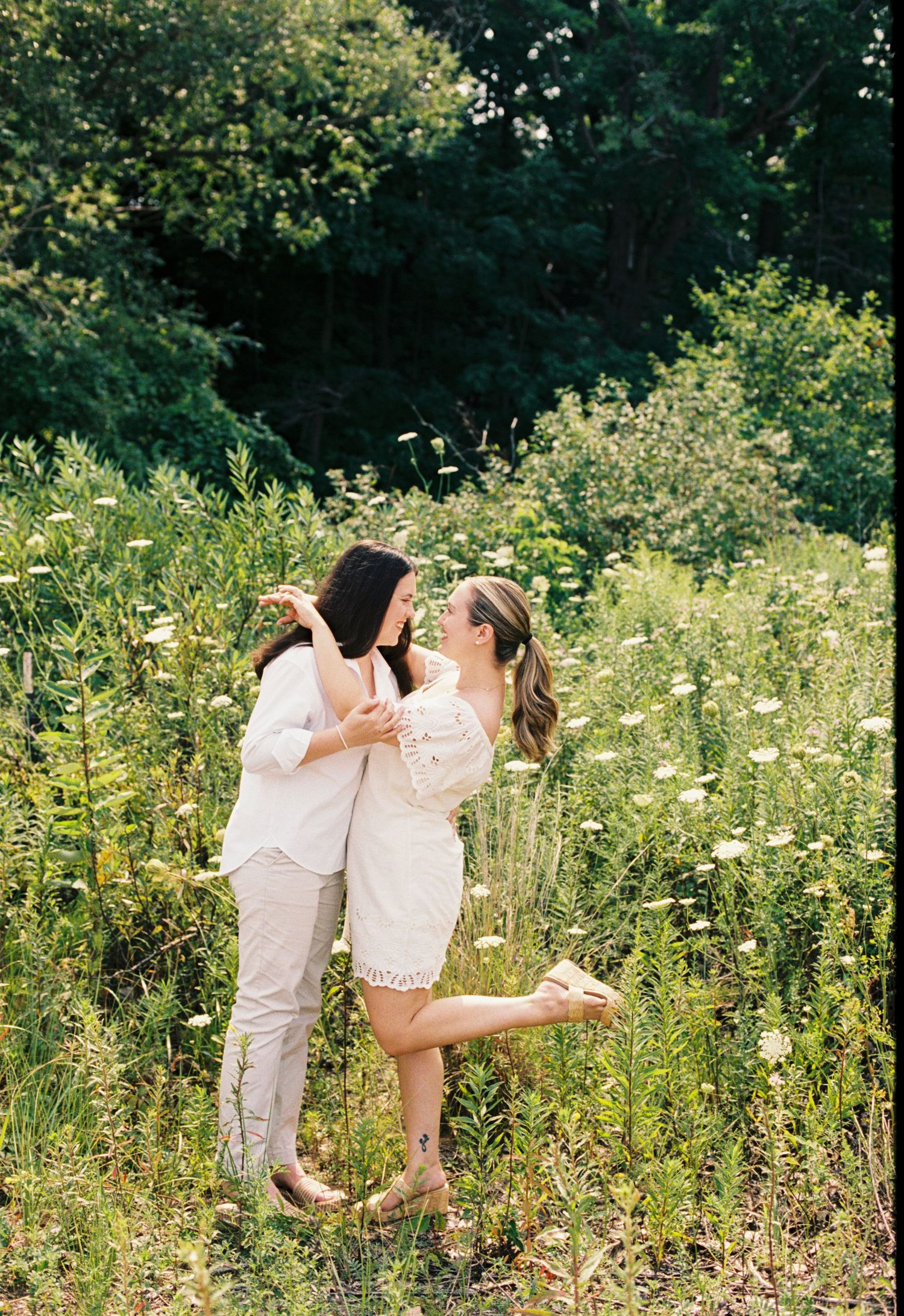 Two women in white outfits laugh and dance together in a lush wildflower meadow during an engagement photo session.