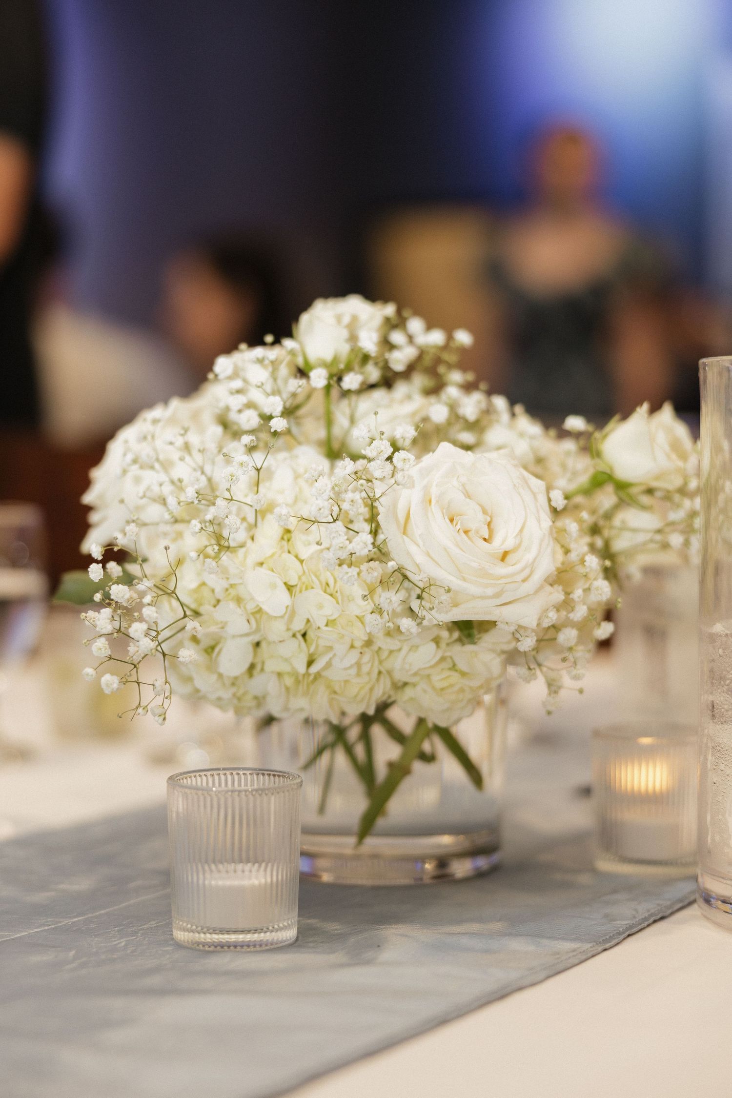 Elegant white wedding centerpiece with roses, hydrangeas, and baby's breath in a glass vase, flanked by votive candles.