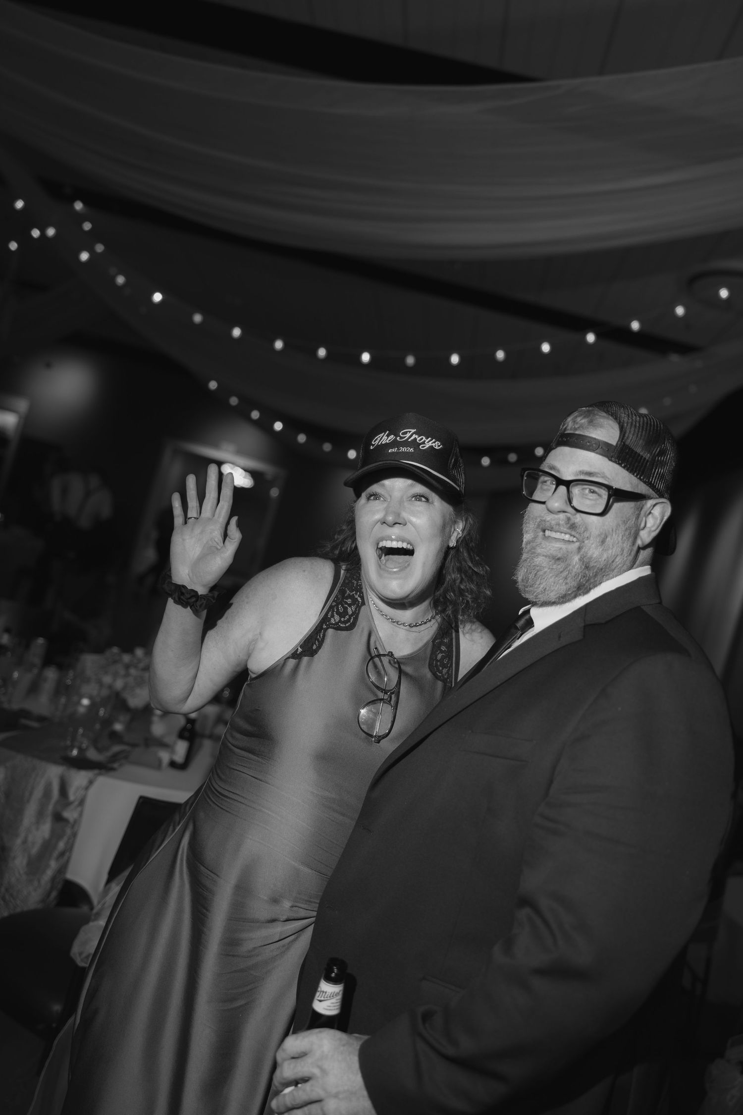 Black and white photo of a laughing woman in a cap and dress waving beside a bearded man in glasses at an indoor event.