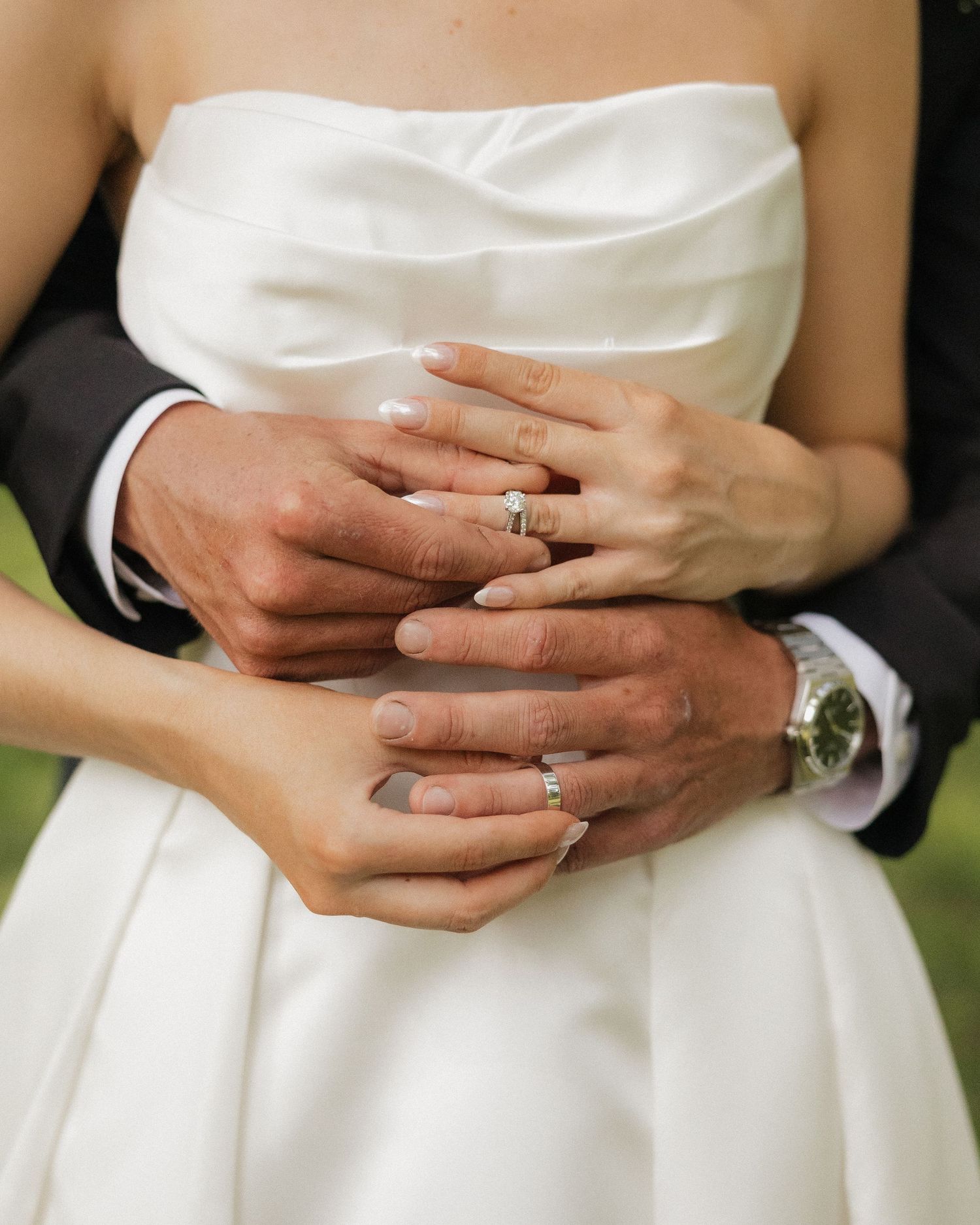 Bride and groom displaying wedding rings, hands intertwined over white bridal gown, groom wearing watch.