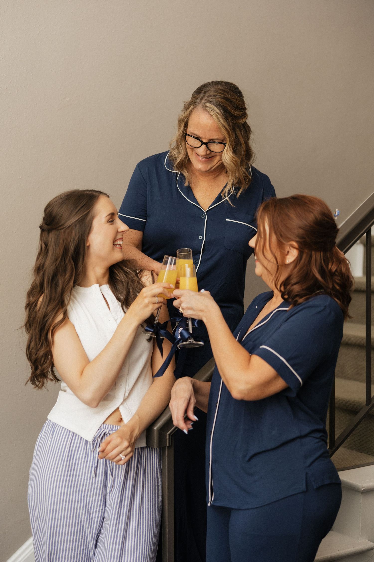 Three women toasting with mimosas on a staircase, two wearing navy pajamas and one in a white top.
