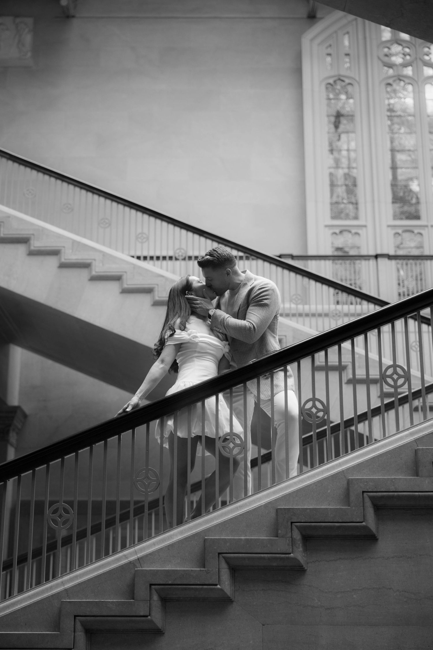 Black and white photo of a couple sharing a romantic kiss on an elegant staircase with ornate railings and stained glass.