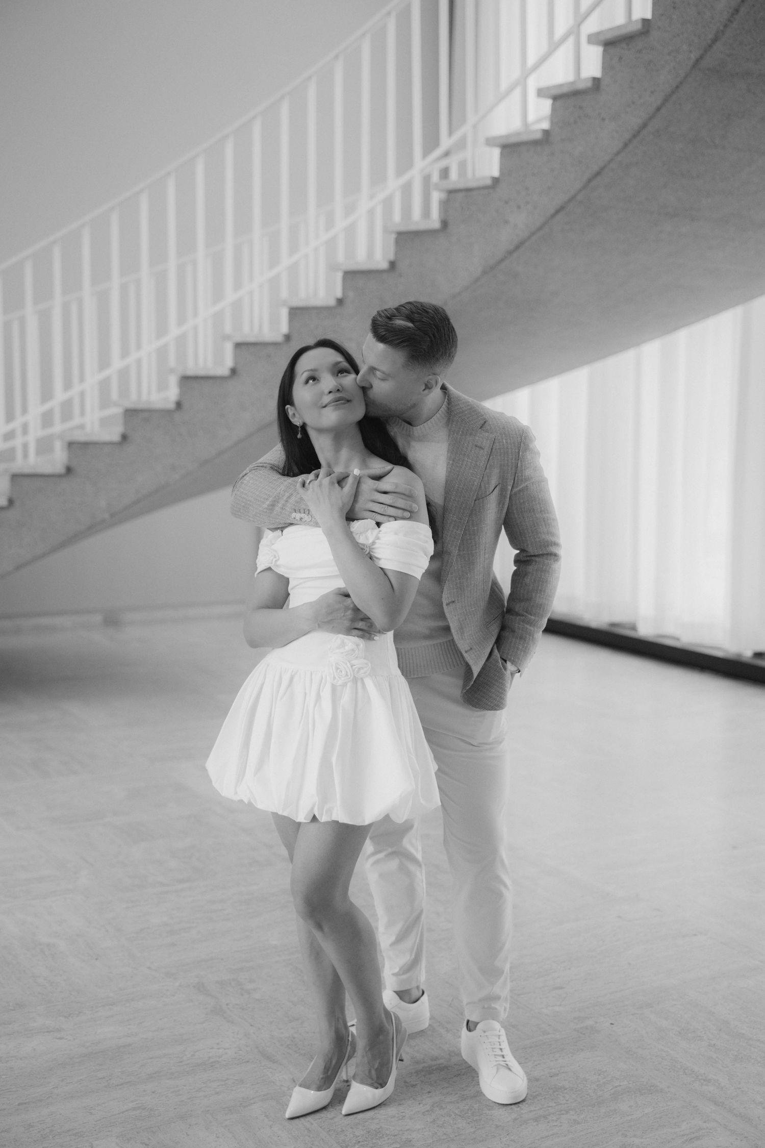 Black and white photo of couple embracing by spiral staircase, woman in white dress, man in suit.